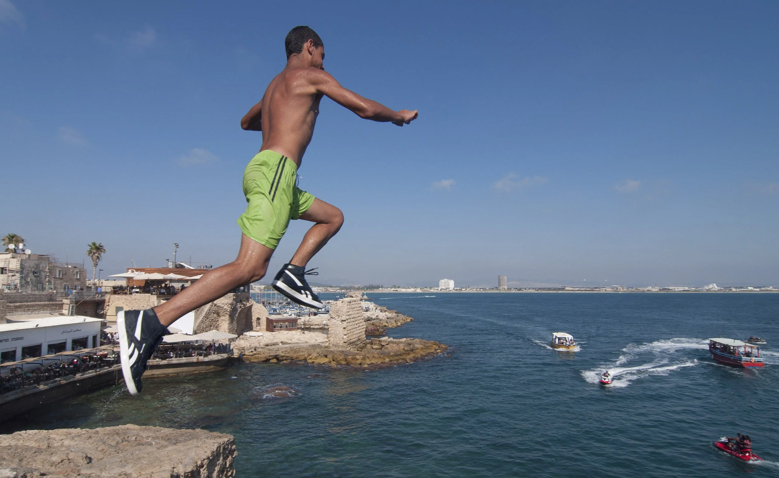 Un joven saltando desde las murallas de San Juan de Acre hacia el mar, con varias embarcaciones navegando en el agua.