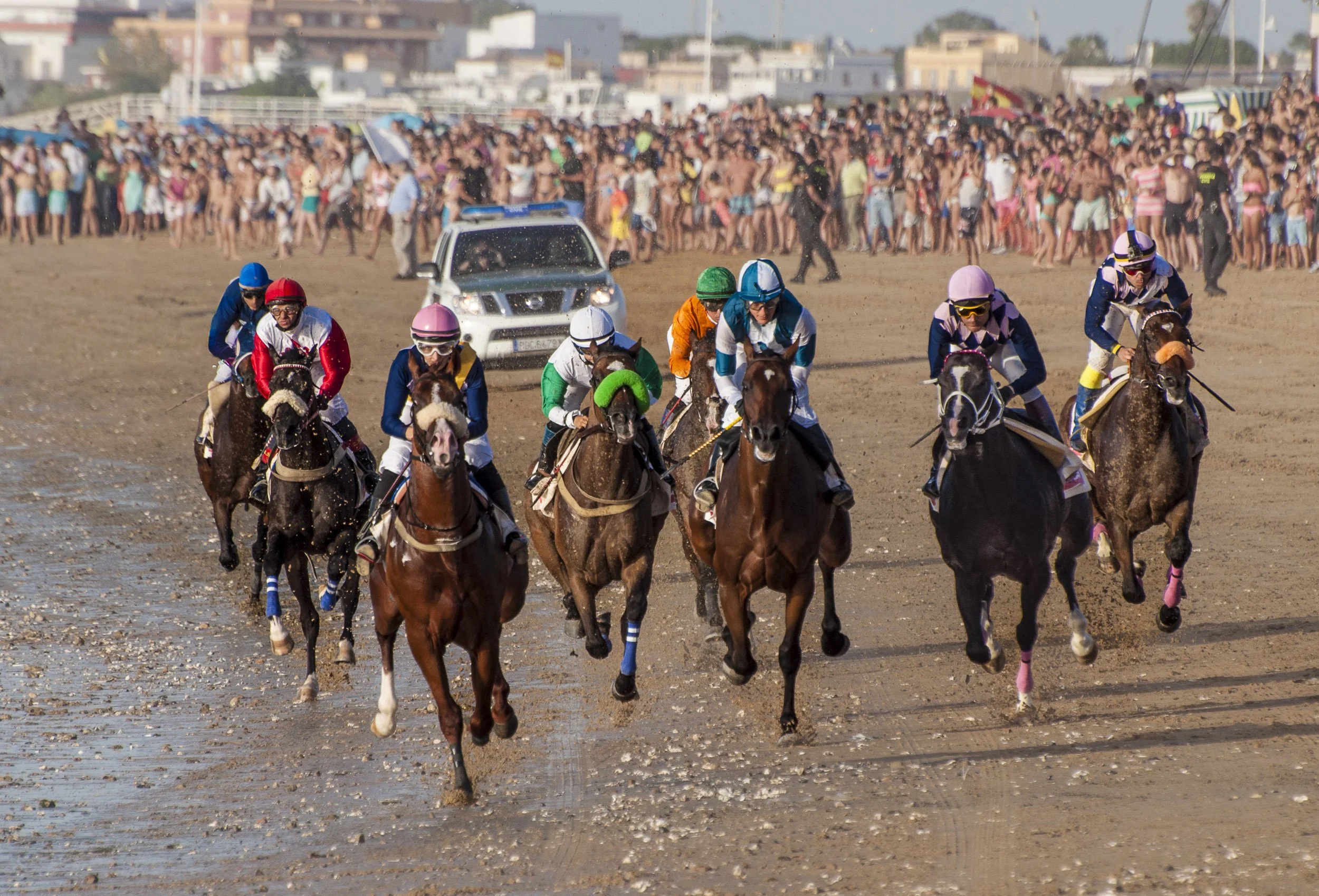 Carrera de caballos en la playa de Sanlúcar de Barrameda, con una gran multitud de espectadores en el fondo.