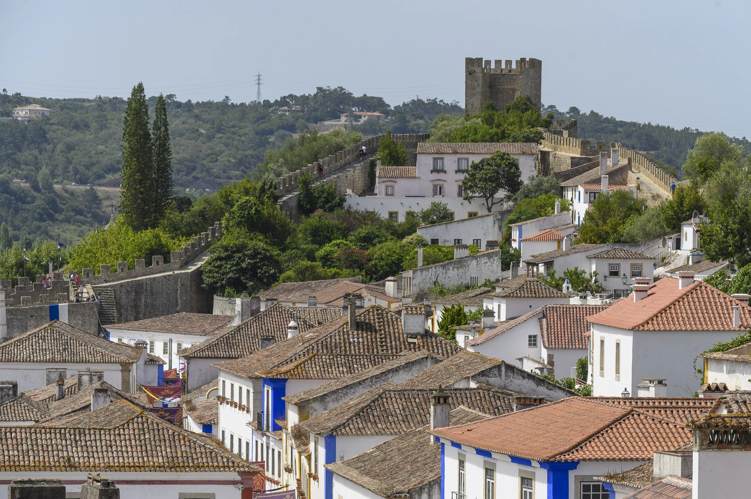 Vista de Óbidos, en Portugal, con casas blancas y tejados de teja roja, rodeada de vegetación y una muralla antigua en la colina con una torre al fondo