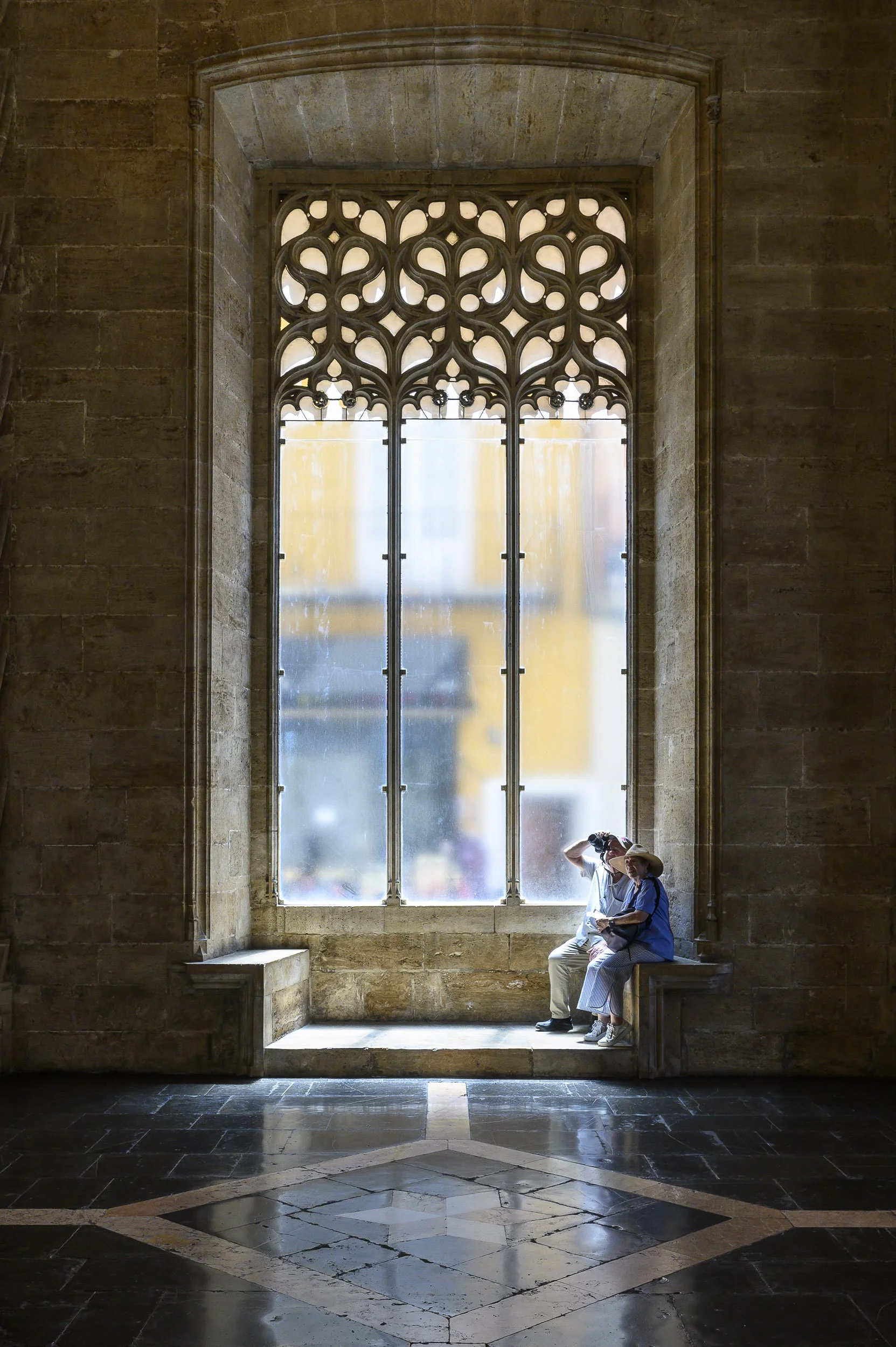 Dos personas sentadas en un banca junto a una gran ventana con vitrales en una catedral, siendo iluminadas por la luz del exterior.