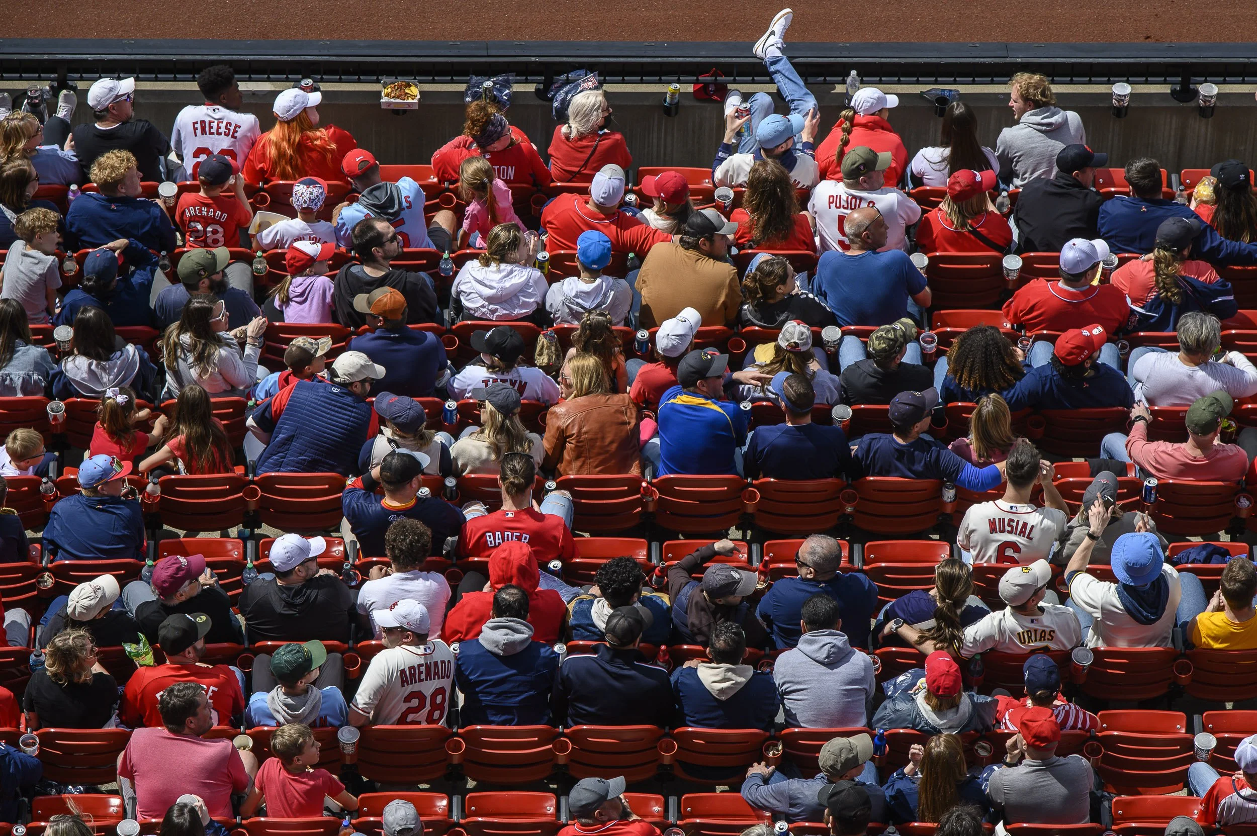 Asistentes en gradería de estadio de béisbol con camisetas y gorras de diversos colores, viendo un partido.