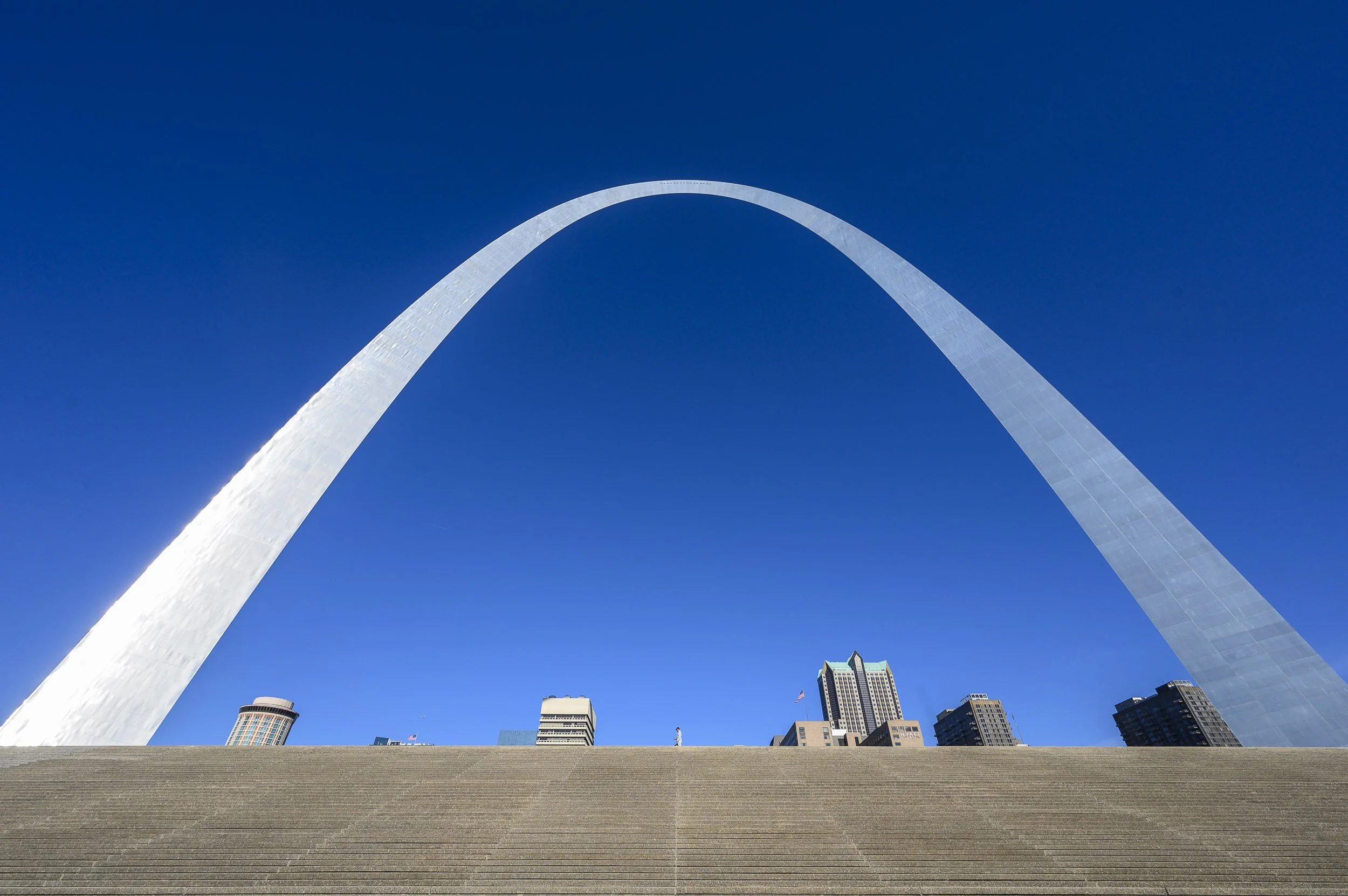 Vista de la Gateway Arch de St. Louis bajo un cielo claro, con edificios altos al fondo y escalones en primer plano.