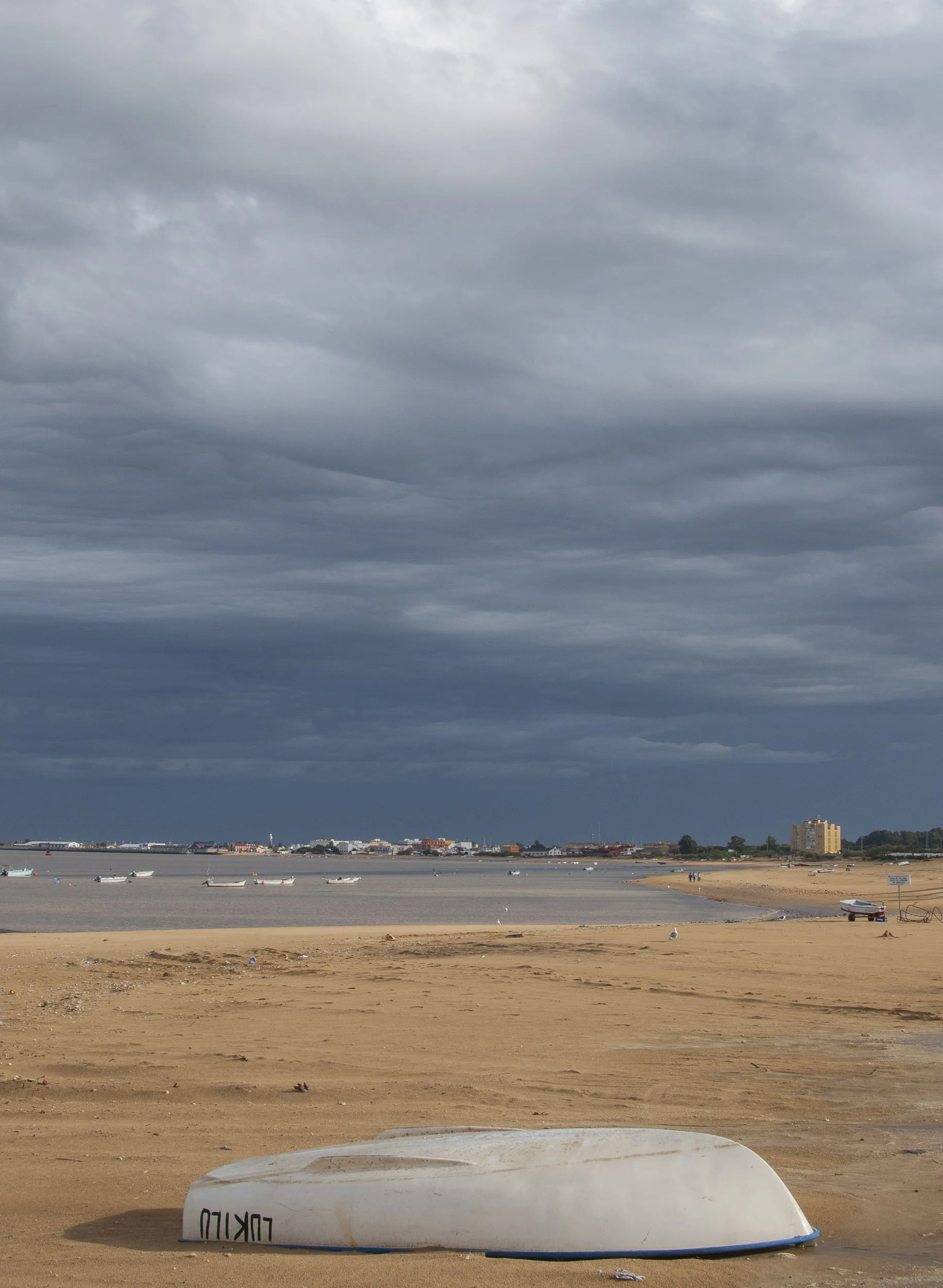 Playa con barco volteado en la arena, barcos en el agua y cielo nublado en un día gris.