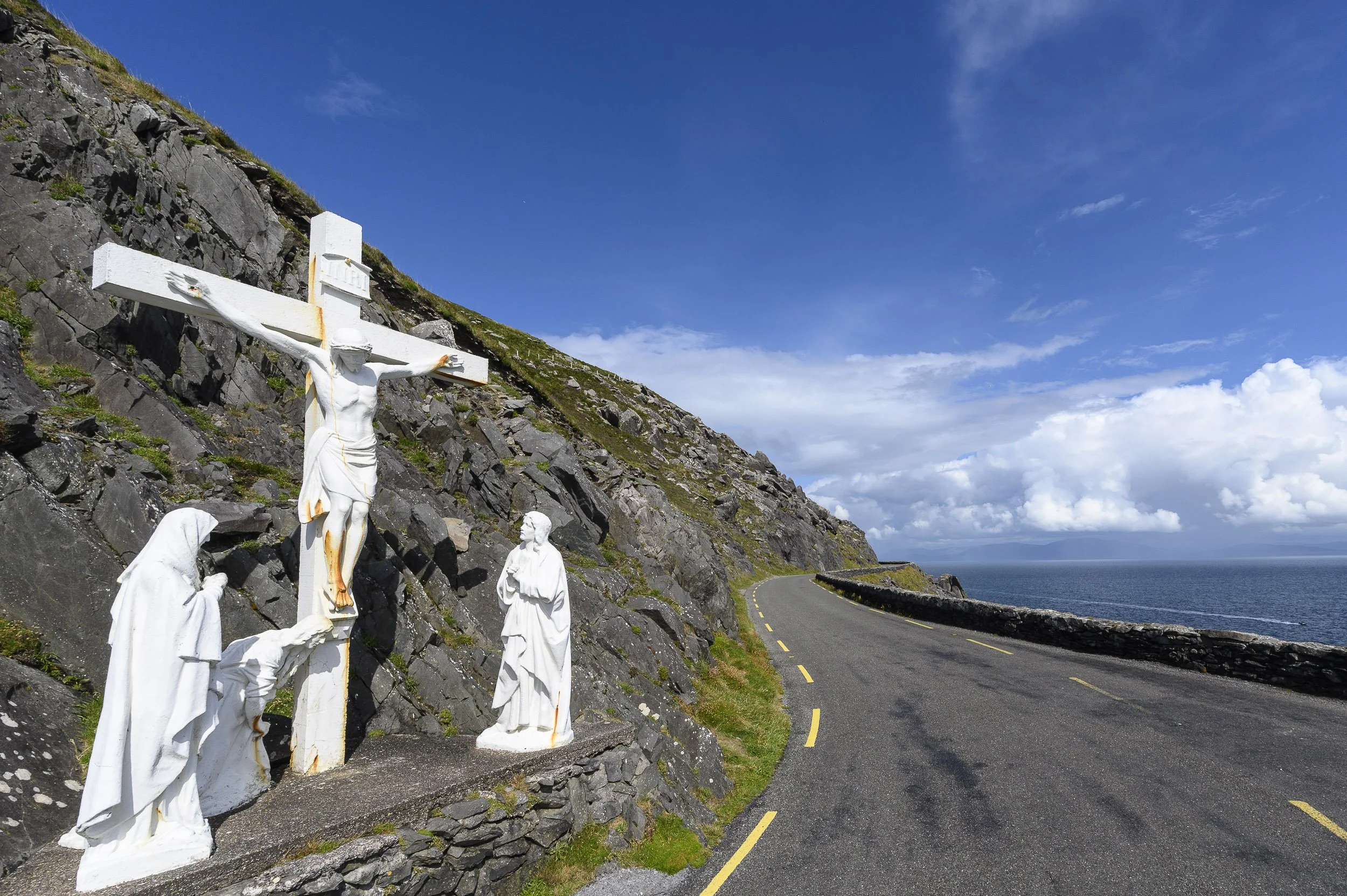 Estatua de Jesucristo crucificado junto a figuras religiosas en una orilla junto a una carretera cercana al mar, en un paisaje costero con cielo azul y nubes en la península de Dingle, Irlanda.
