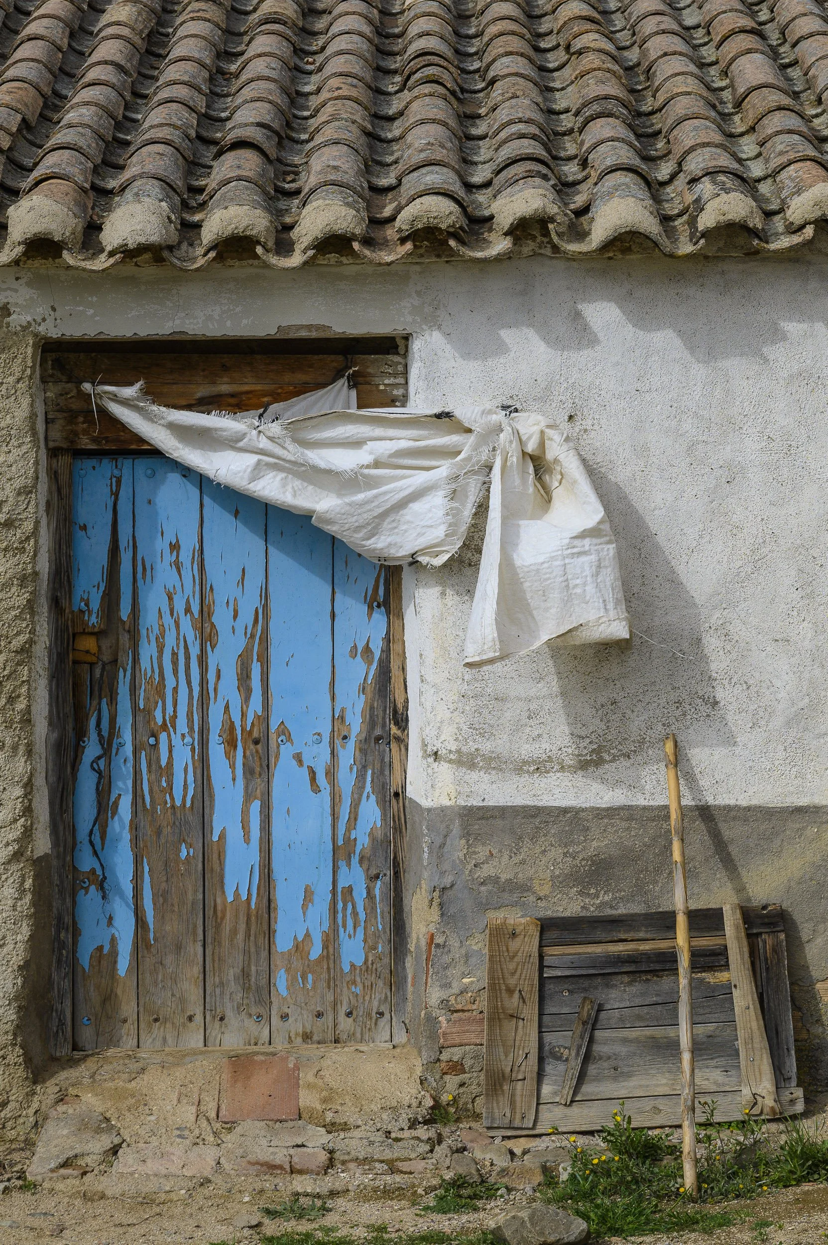 Puerta de madera vieja pintada de azul, con tela blanca colgando, en una pared de cemento. Interior de tejas de cerámica en el techo, y madera apilada a la derecha. Tierra y piedras en el suelo con plantas pequeñas.