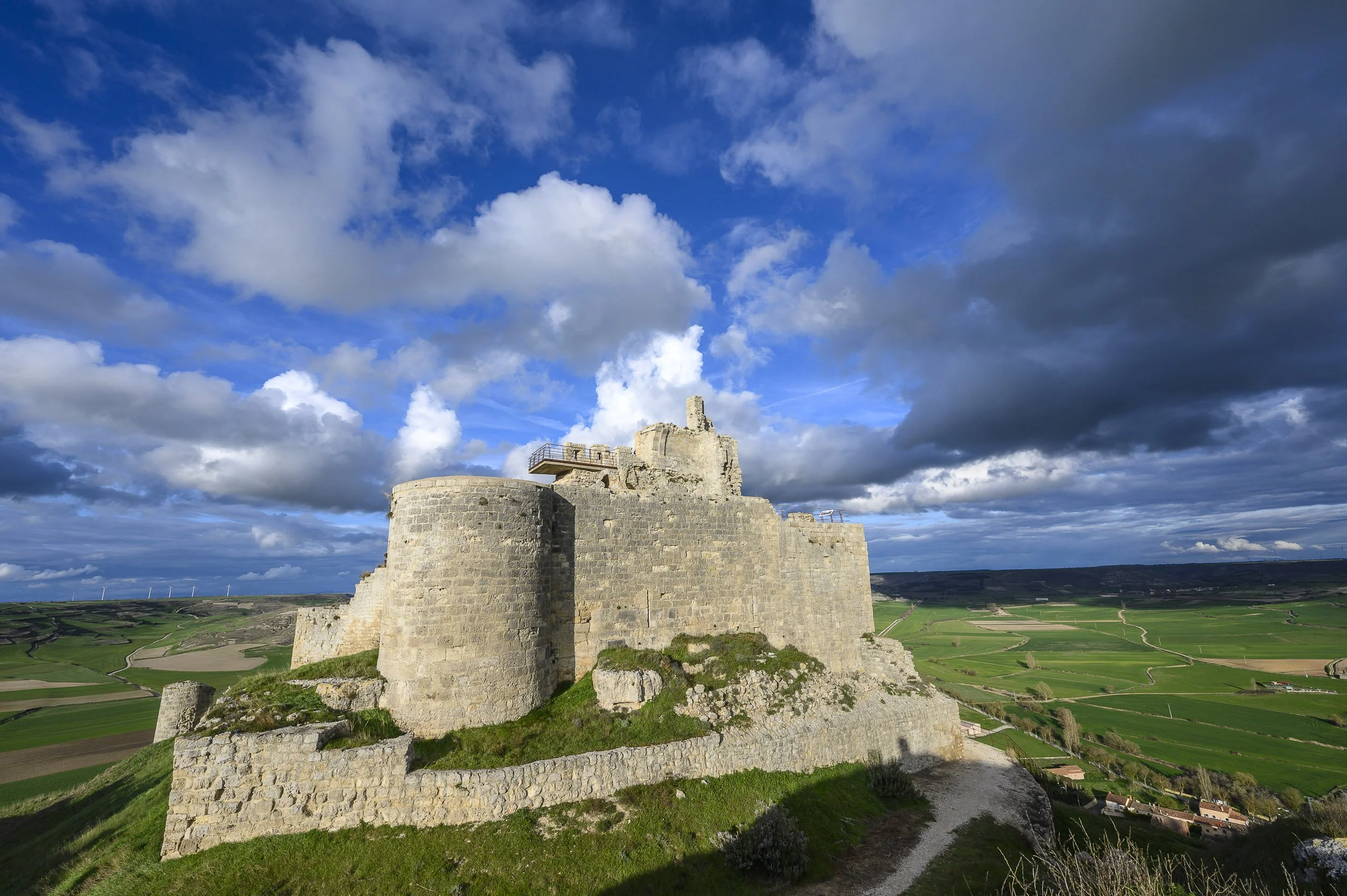 Castillo de Castrojériz en una colina rodeado de campos verdes y un cielo con nubes.