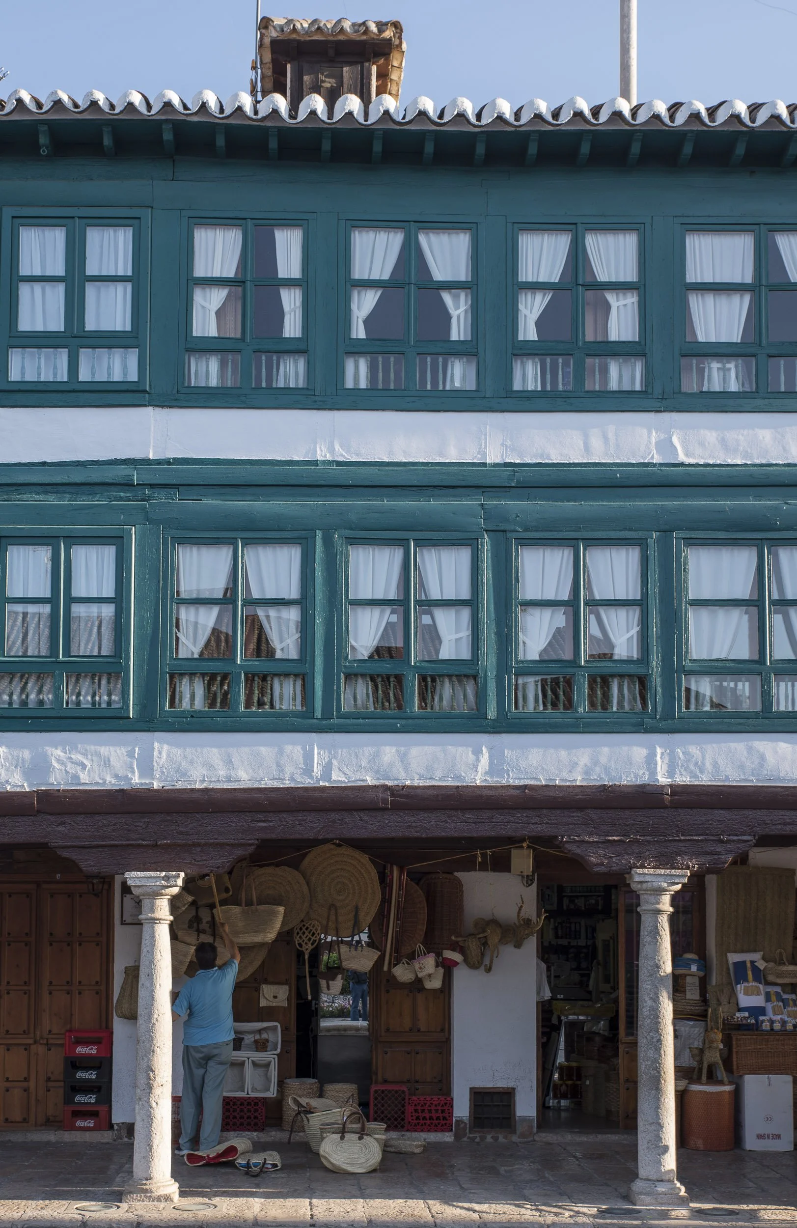 Detalle de la Plaza Mayor de Almagro, con ventanas de madera pintadas de azul y cortinas blancas, en la planta baja un puesto con artículos de mimbre y una persona viendo el objeto en una de las paredes.
