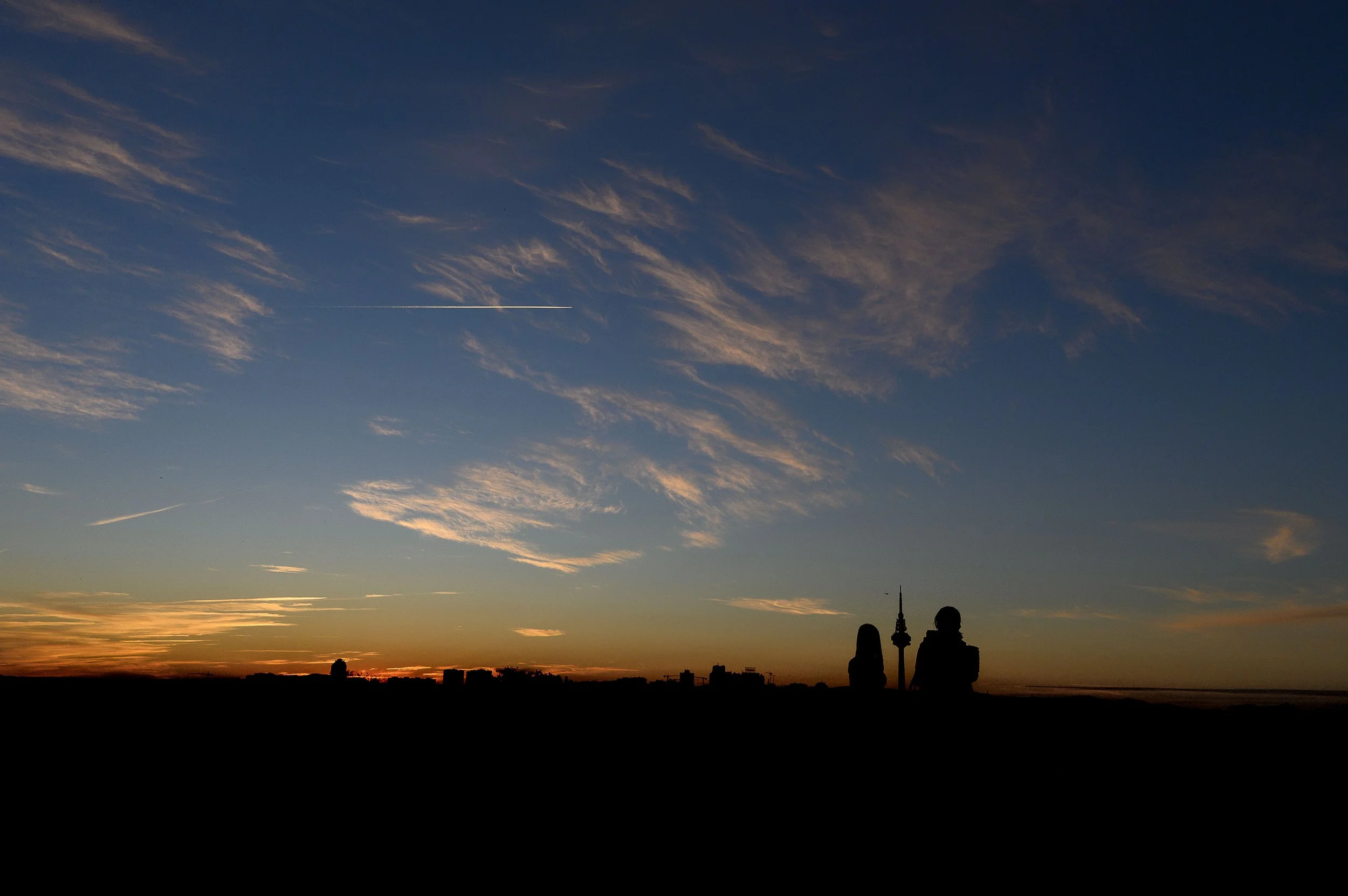 Siluetas de dos personas sentadas en un campo viendo la puesta del sol, con un cielo con nubes y un avión en vuelo.