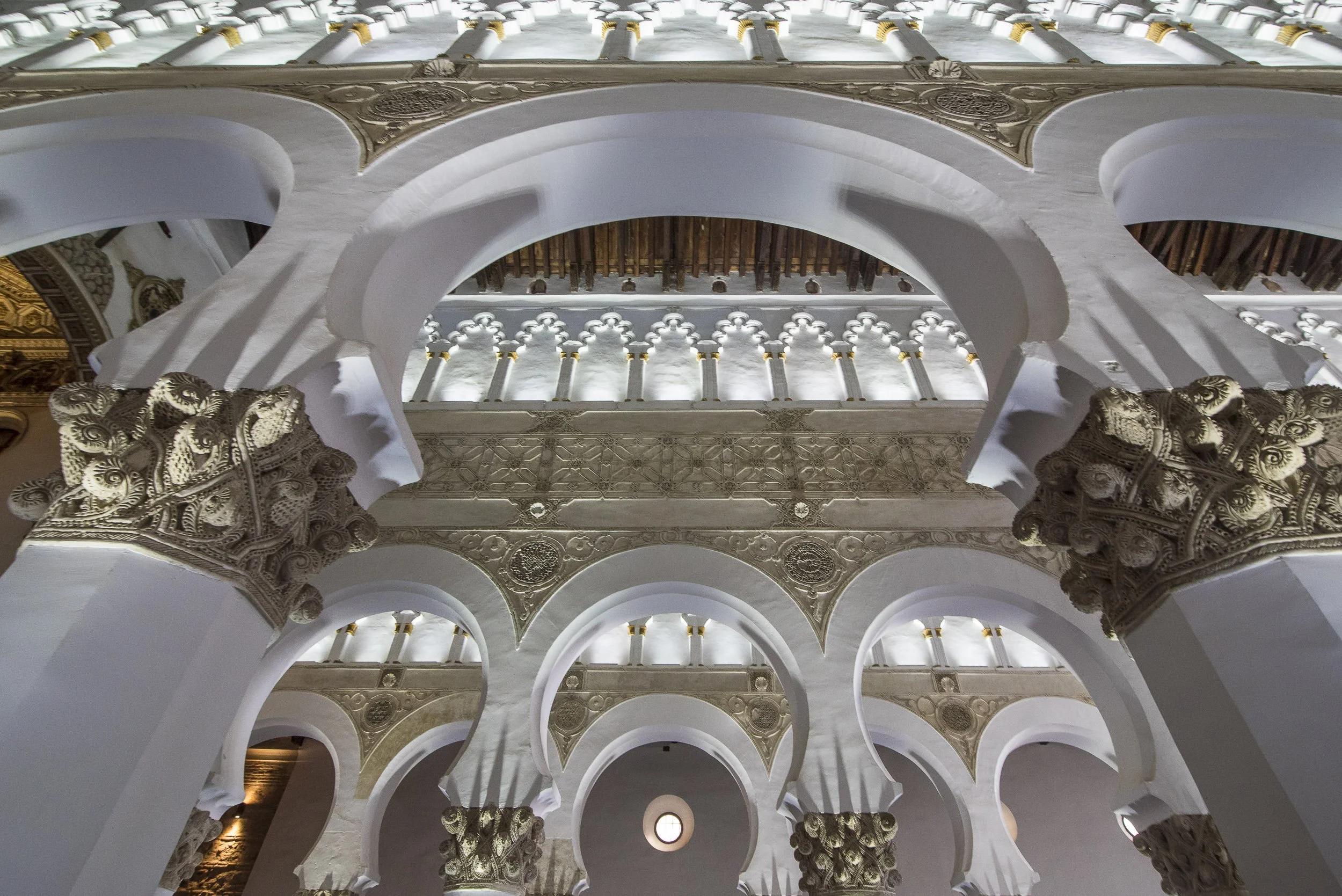 Vista del interior de un edificio con arquitectura colonial, con arcos blancos y detalles ornamentales dorados y esculturas en las columnas.