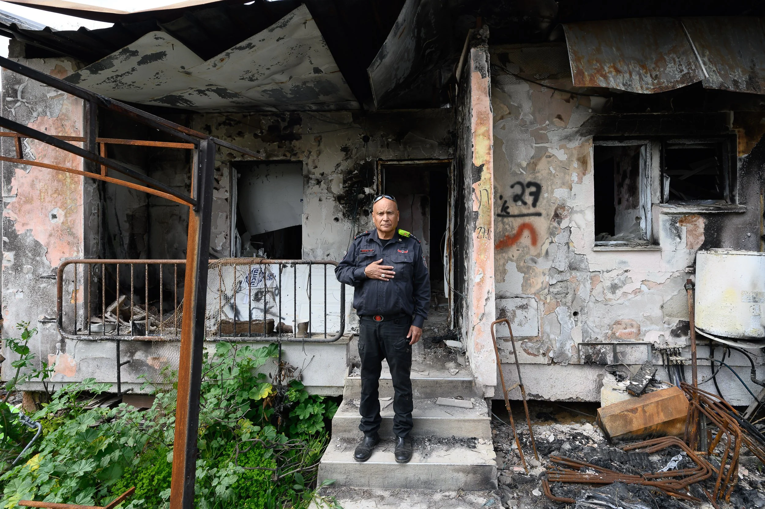 Un oficial de policía frente a una casa quemada y dañada por un incendio, con paredes calcinadas y objetos metálicos quemados a su alrededor.