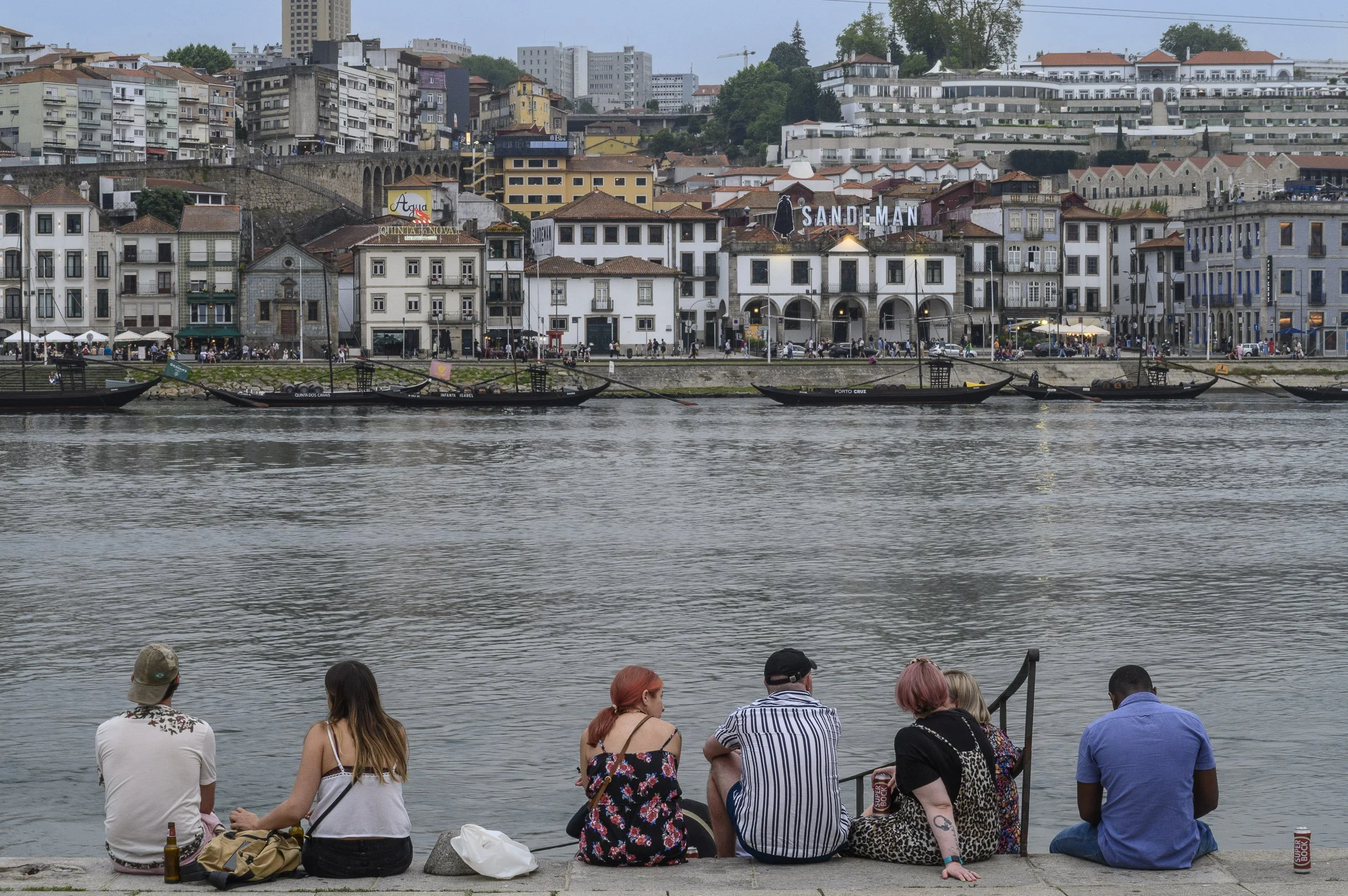 Personas sentadas junto al río Duero en Oporto, en un día nublado, con edificios y barcos tradicionales al fondo.