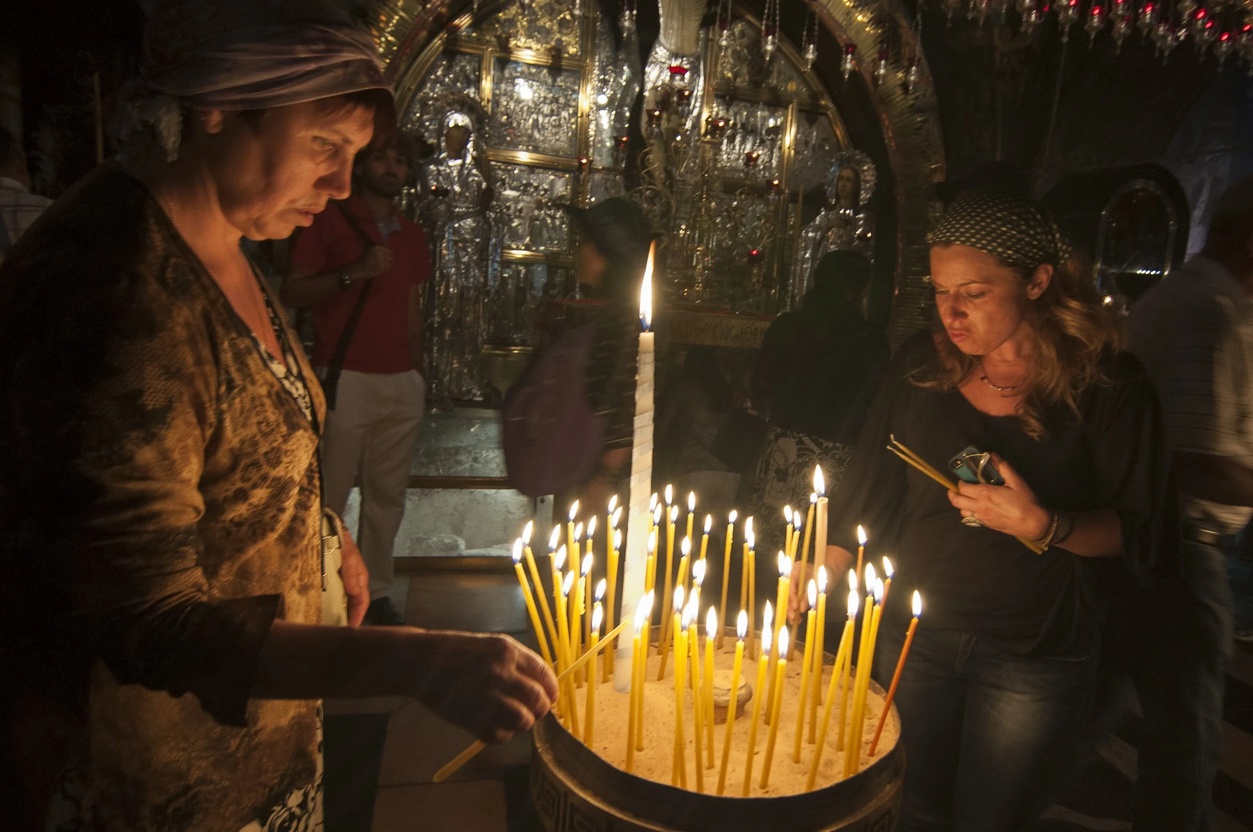 Personas encendiendo velas dentro de la iglesia del Santo Sepulcro, en Jerusalén, con altares decorados con objetos religiosos y estatuas en el fondo.