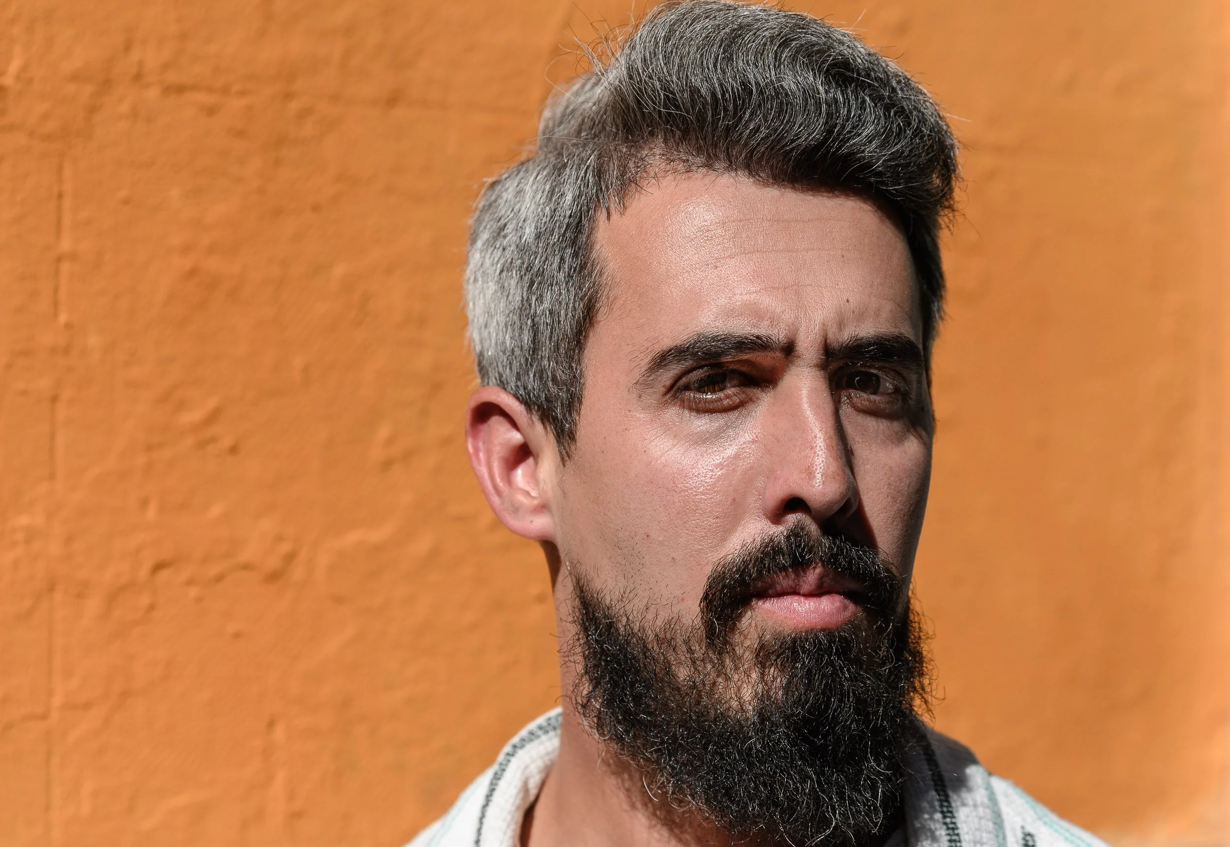 Hombre con barba y cabello gris, con expresión seria, frente a fondo naranja.