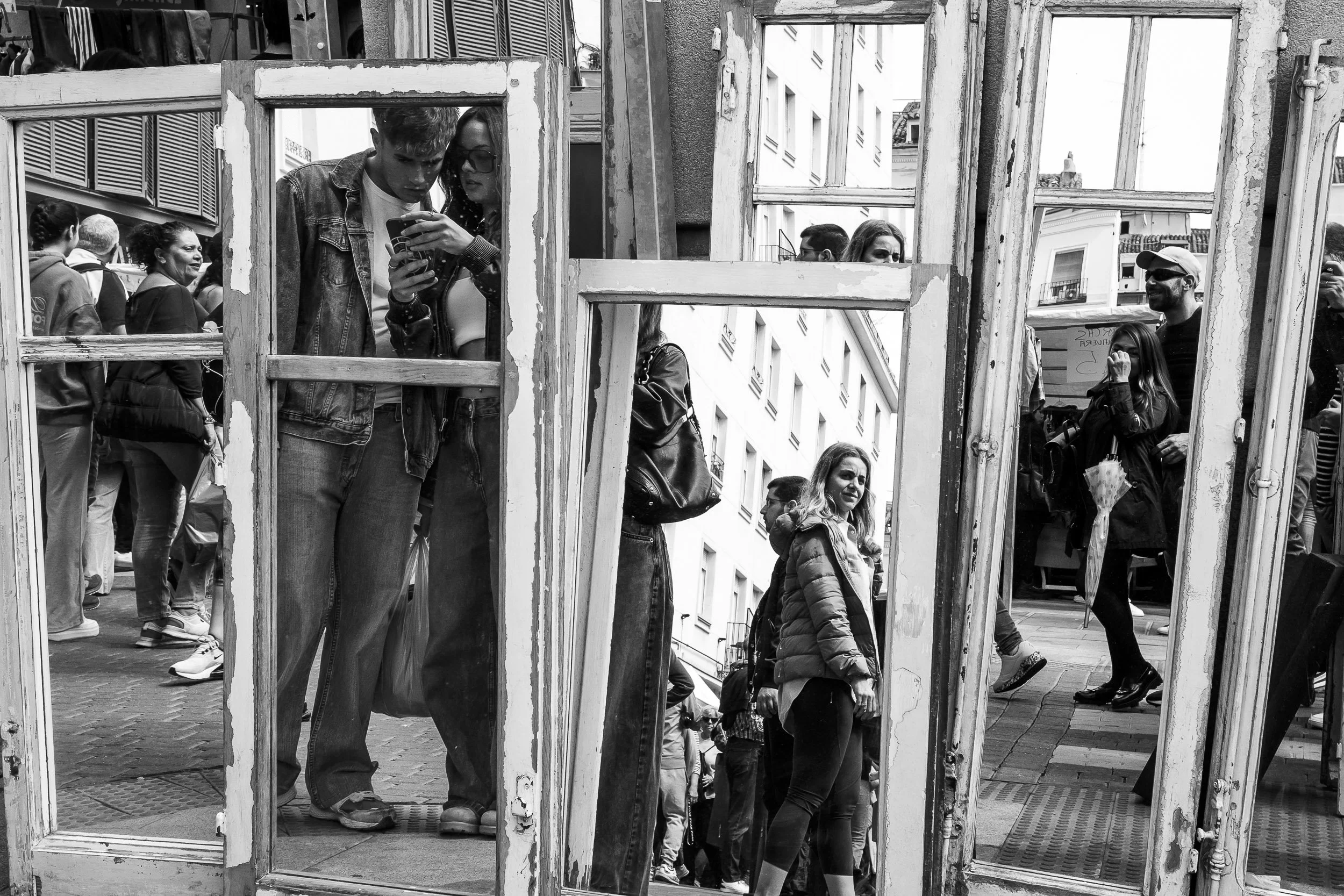 Variedad de personas en una calle, vistas a través de una estructura de madera envejecida con múltiples ventanas. Algunas personas observan, otras caminan, y algunas parecen interactuar, en un ambiente urbano.