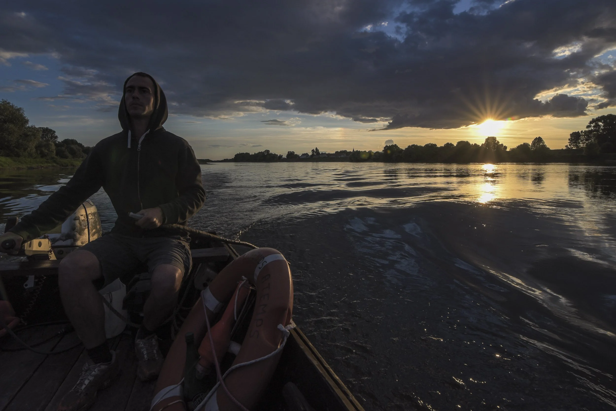 Joven en barco navegando por un río al atardecer, con cielo nublado y sol en el horizonte.