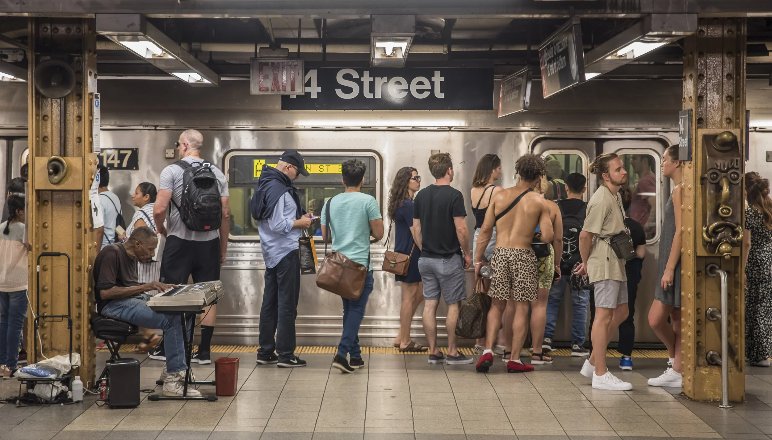Gente en la estación de metro en la línea 4, esperando para abordar el tren, algunos leen, otros conversan y un músico toca un teclado a la izquierda.