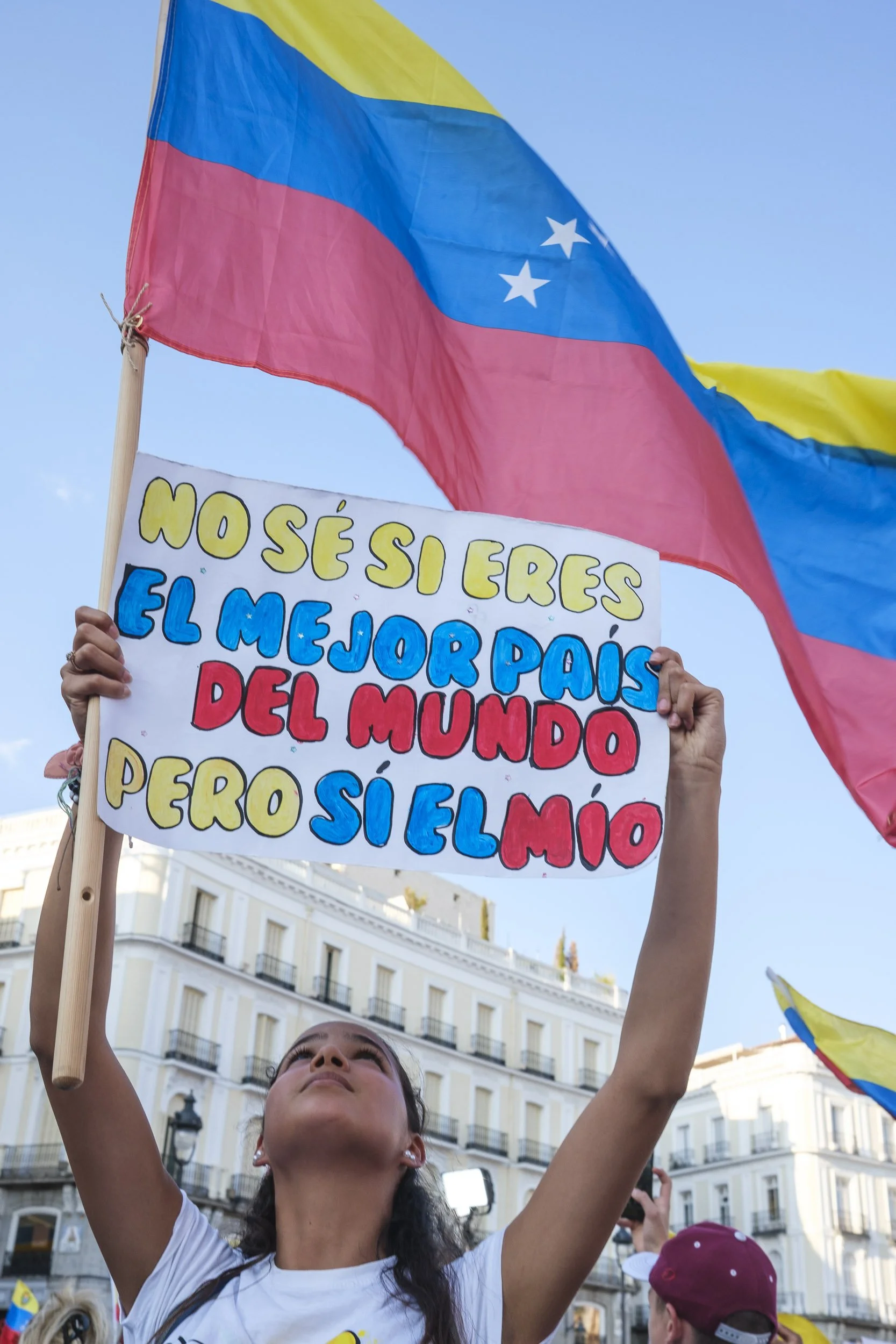 Joven sosteniendo cartel con texto en español y banderas de Venezuela durante una manifestación en una calle de ciudad con edificios antiguos.