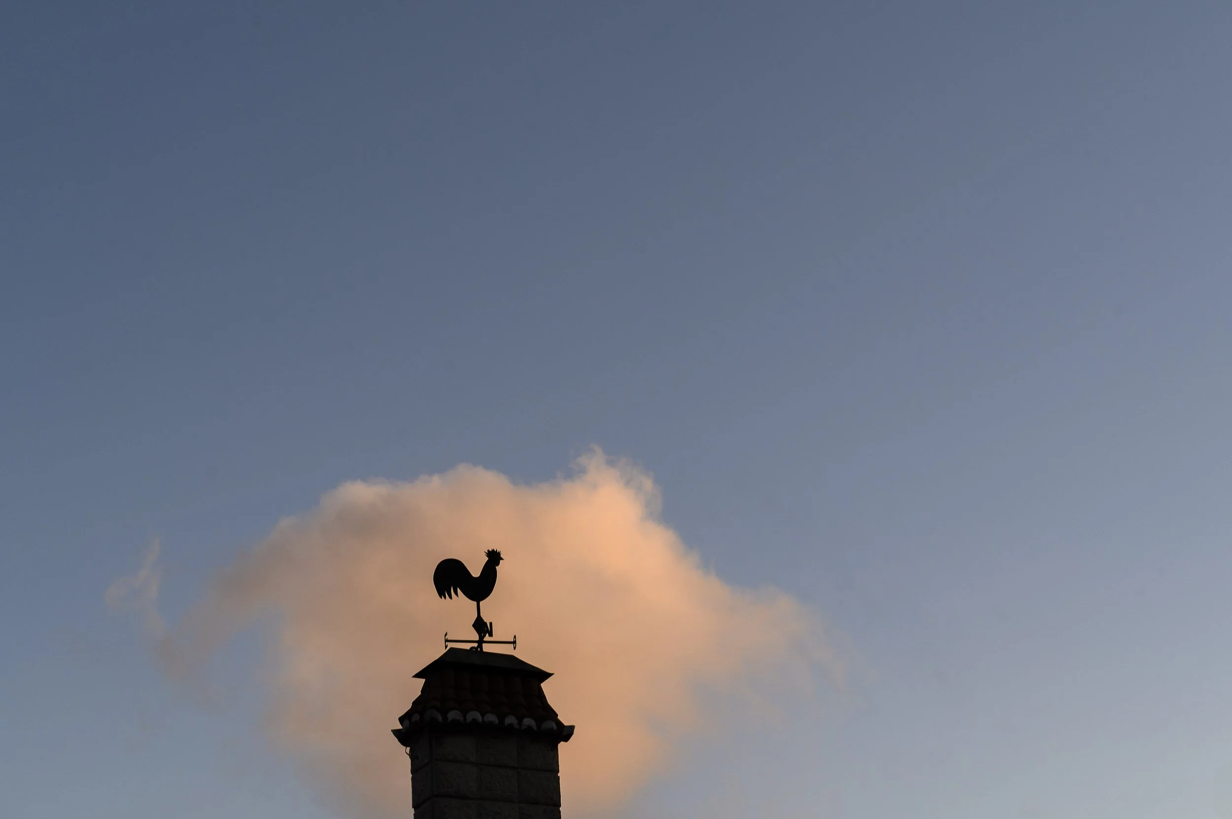 Silueta de un gallo en la veleta en la cima de una iglesia de Lisboa, con cielo azul y una nube al fondo.