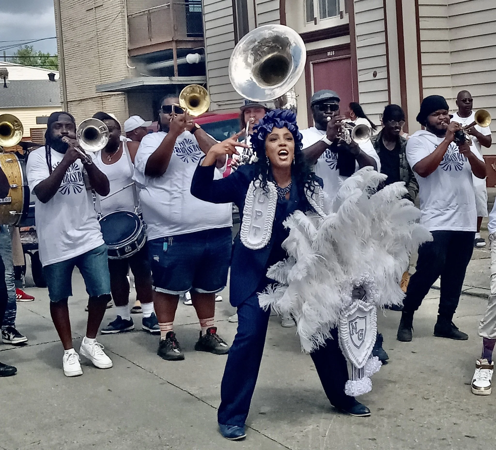 Group of musicians playing brass and drums in a street parade, with a woman in a costume with feathers and a blue headpiece in front.