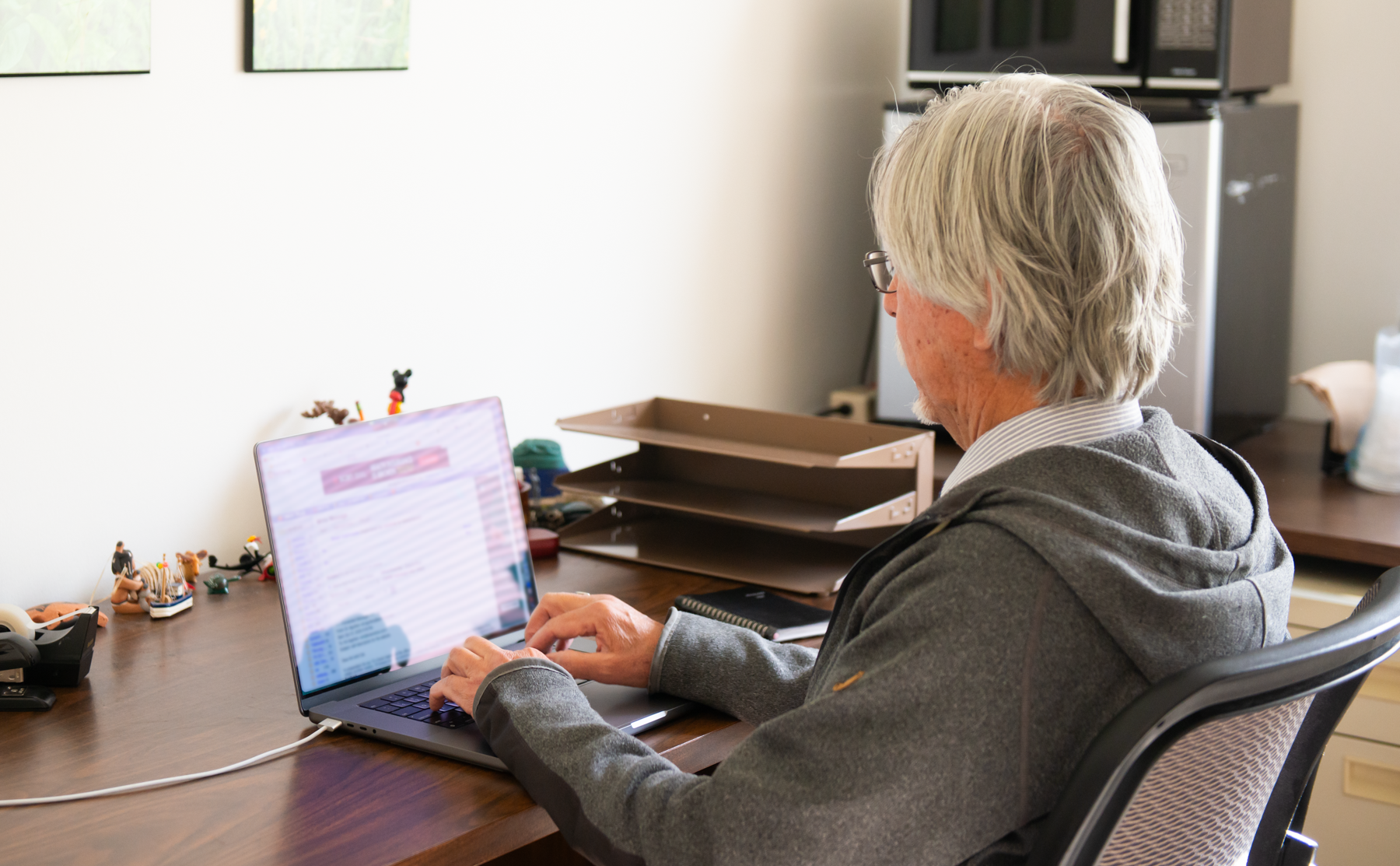 An elderly man with gray hair and glasses sitting at a desk, using a laptop surrounded by office supplies and figurines, with a white wall and microwave oven in the background.