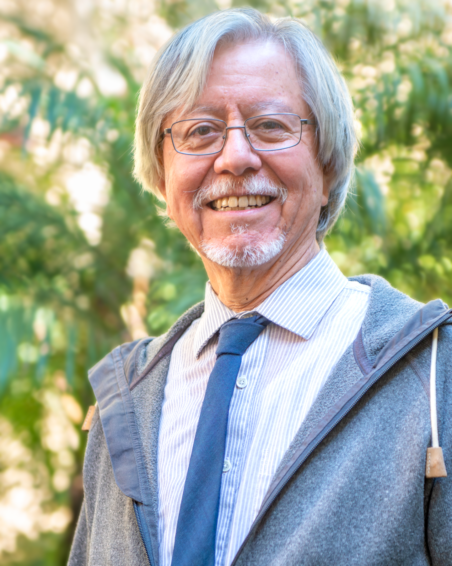 An older man with gray hair, glasses, and a beard smiling outdoors in a park or garden with greenery in the background.