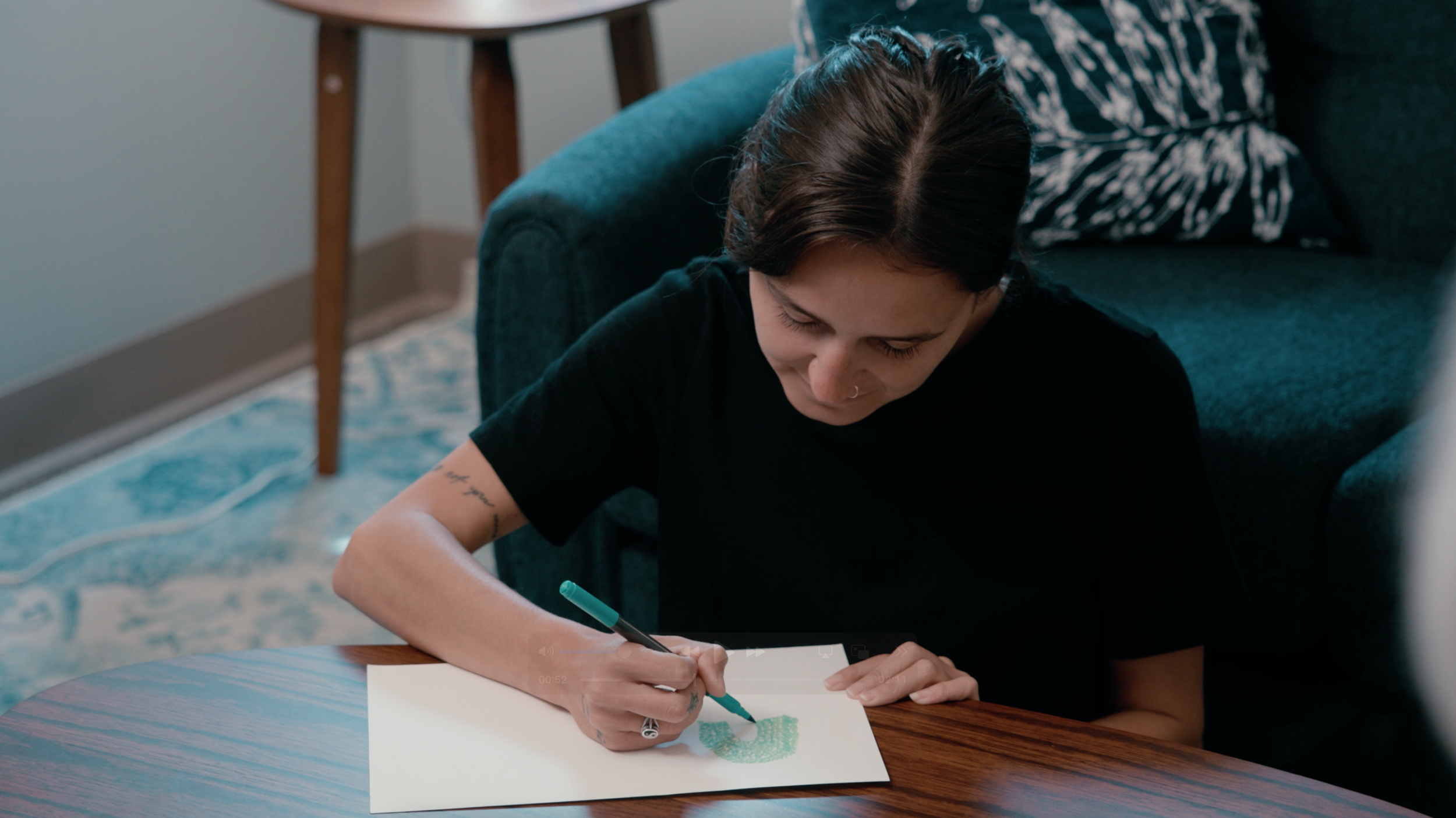 Young woman with dark brown hair, wearing a black t-shirt, drawing a green shape on white paper with a black marker, sitting at a wooden table in a cozy room with a blue carpet and a dark blue sofa in the background.