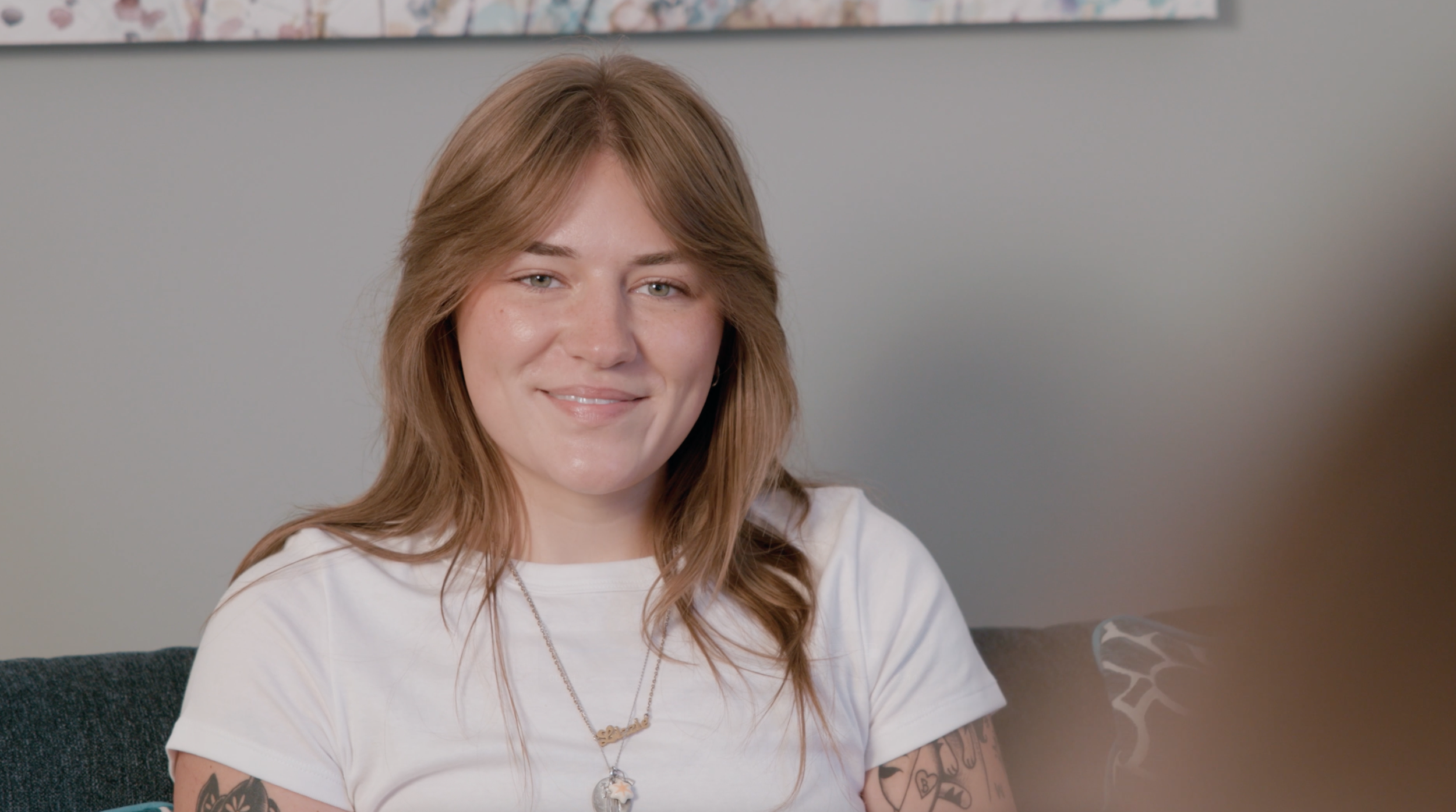 A young woman with light brown hair and tattoos on her arms, wearing a white T-shirt and a necklace, smiling while sitting on a dark couch in a room with a light gray wall.