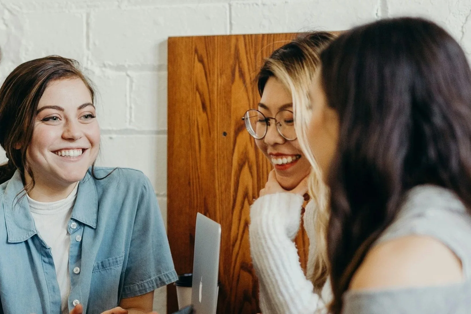 Three young women sitting together in a casual setting, smiling and talking, with one wearing glasses and a white sweater, another in a denim shirt, and the third with dark hair and a gray top.