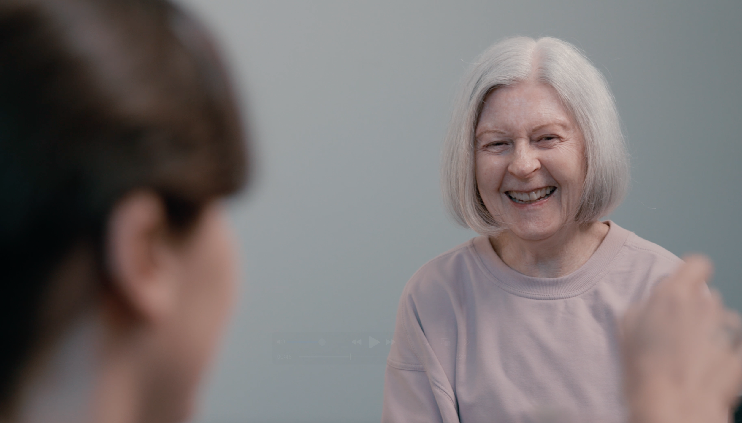 An elderly woman with gray hair smiling and laughing, seen from the front, with someone in the foreground out of focus.