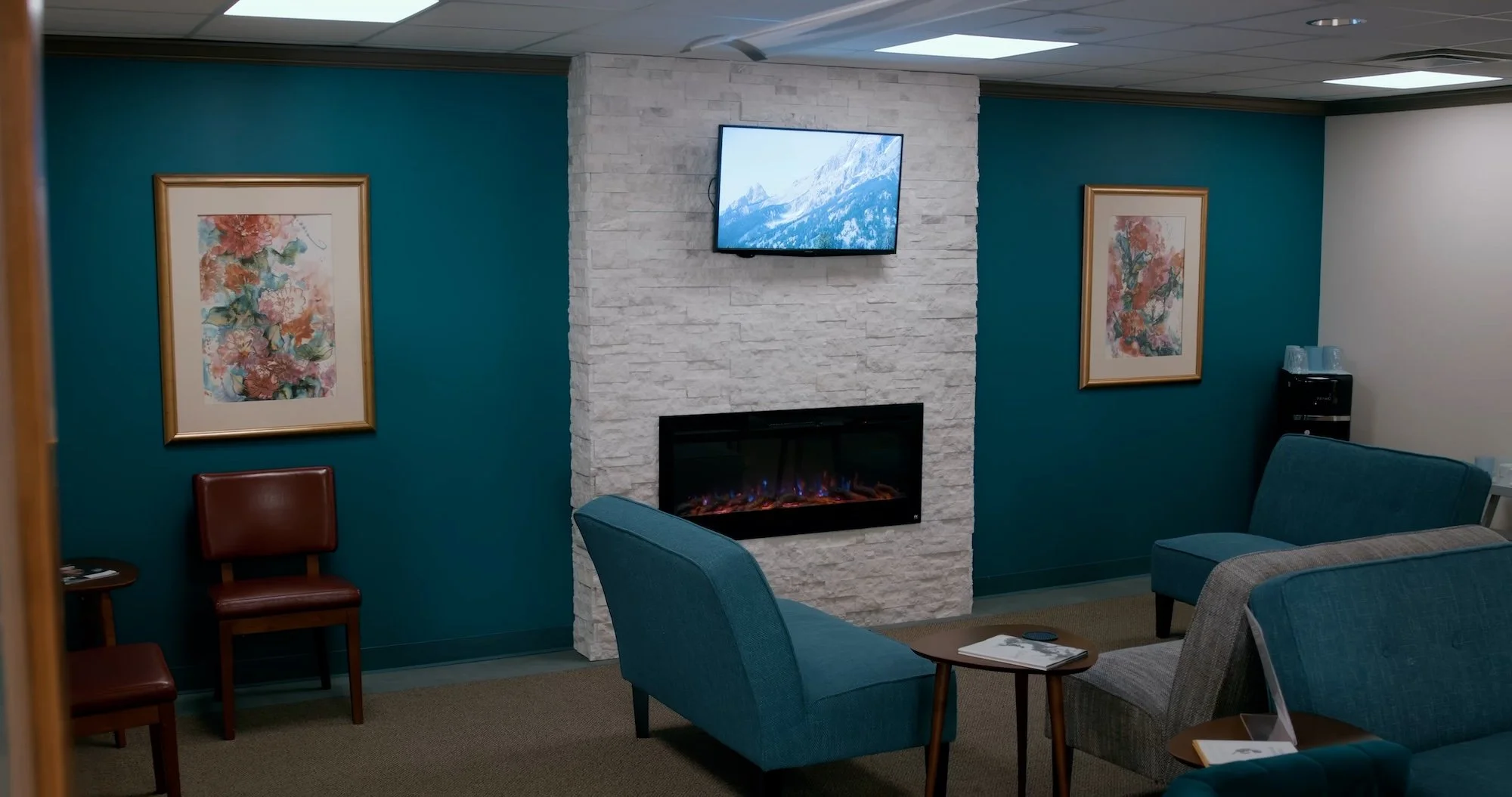 A cozy waiting room with a white stone fireplace, a mounted television displaying mountain scenery, teal walls with framed floral artwork, and various upholstered chairs and side tables.