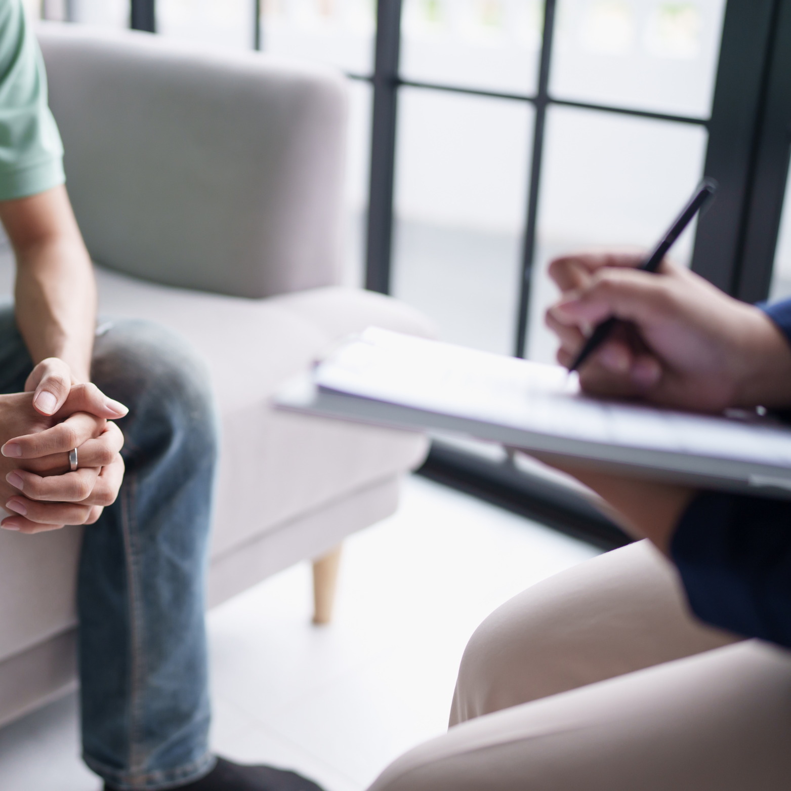 Therapist taking notes during a therapy session with a client sitting nearby, hands clasped on the client's lap.
