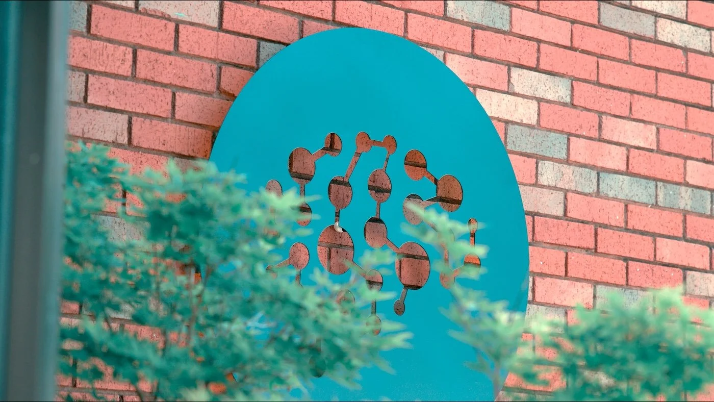 A blue circular sign with cut-out designs of connected circles and lines against a red brick wall, with green bushes in the foreground.