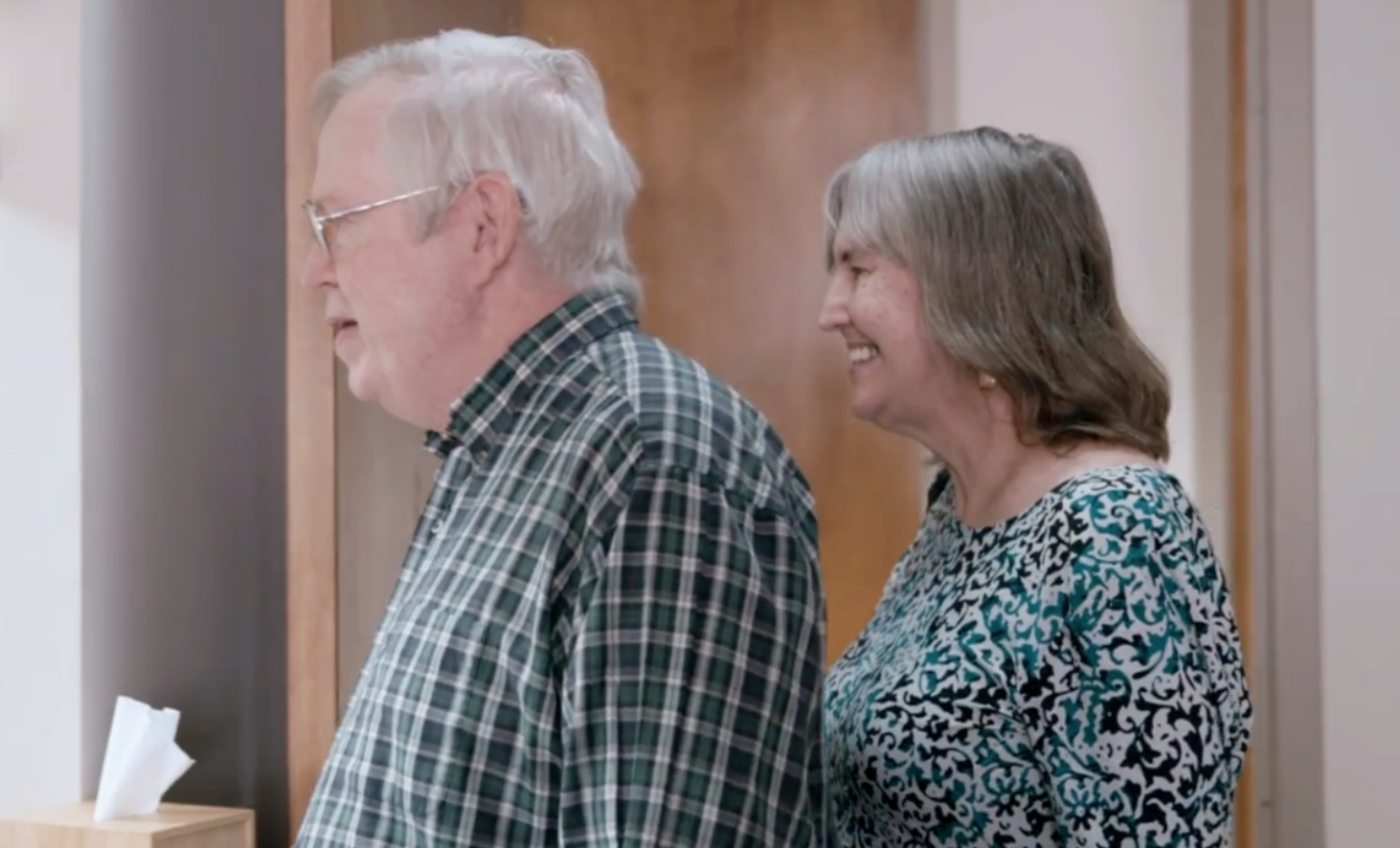 An elderly man with glasses and gray hair standing beside an elderly woman with gray hair. Both are smiling and facing to the left in an indoor setting.