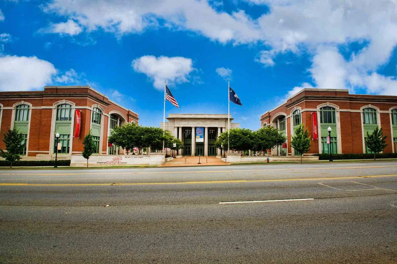 The Chapman Cultural Center building with a wide sidewalk, trees, and flags in front, under a blue sky with clouds.