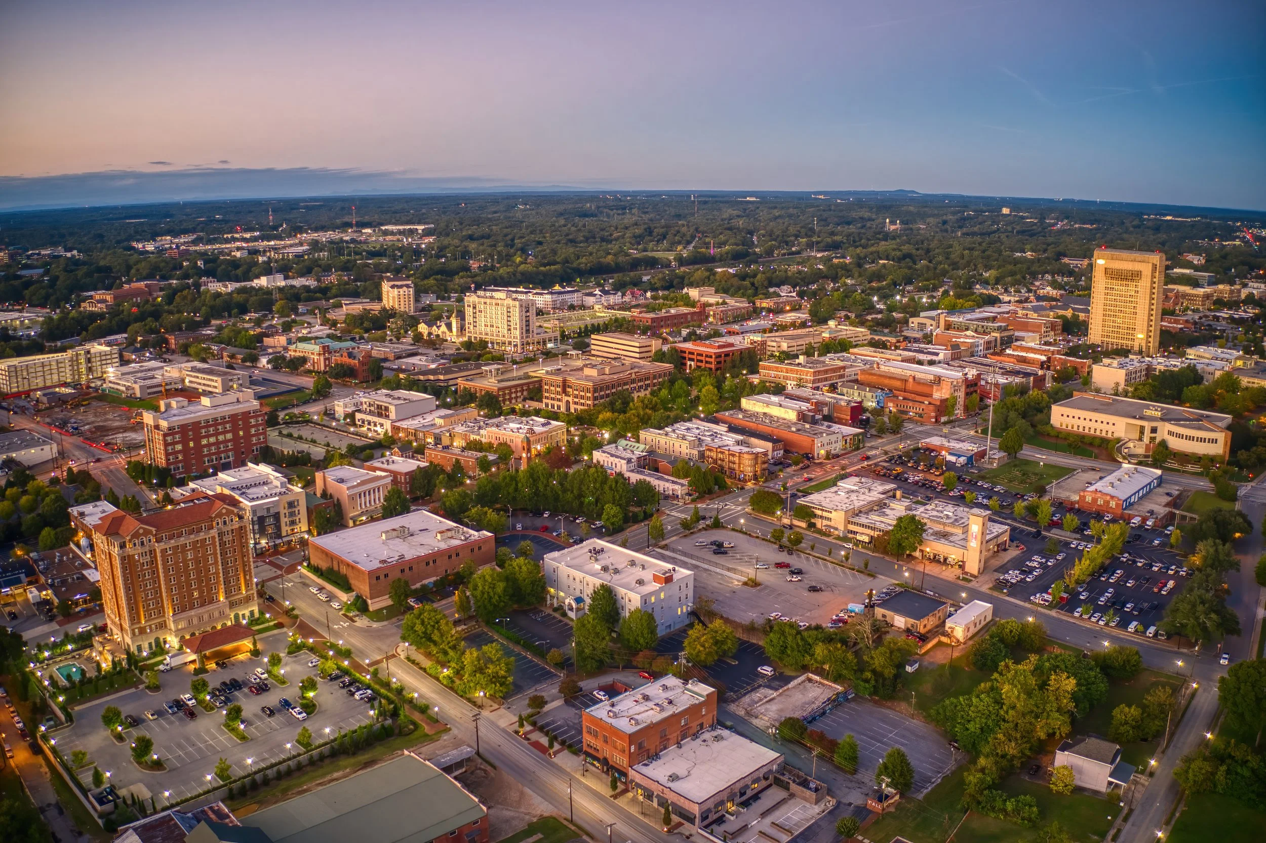 An aerial view of a cityscape at dusk, showing buildings, trees, streets, and parking lots with lighted streets and a horizon with a gradient sky.