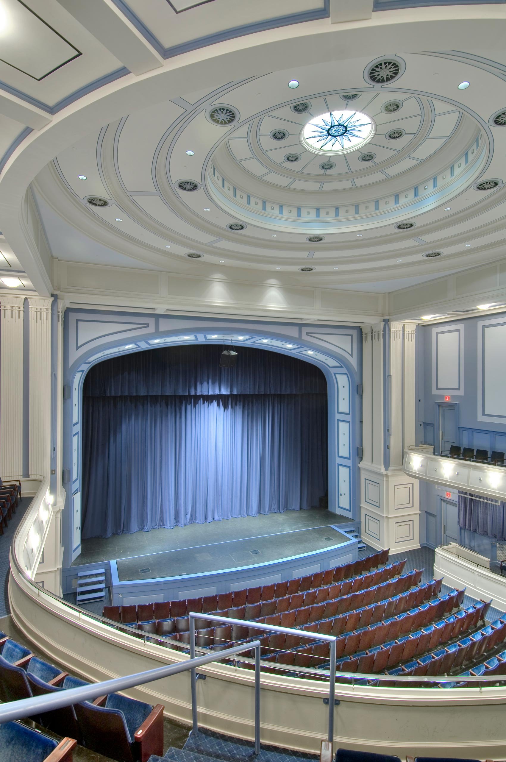 Interior view of an empty theater with a stage and blue curtains, rows of red cushioned seats, and decorative light blue and white walls, taken from the balcony level.