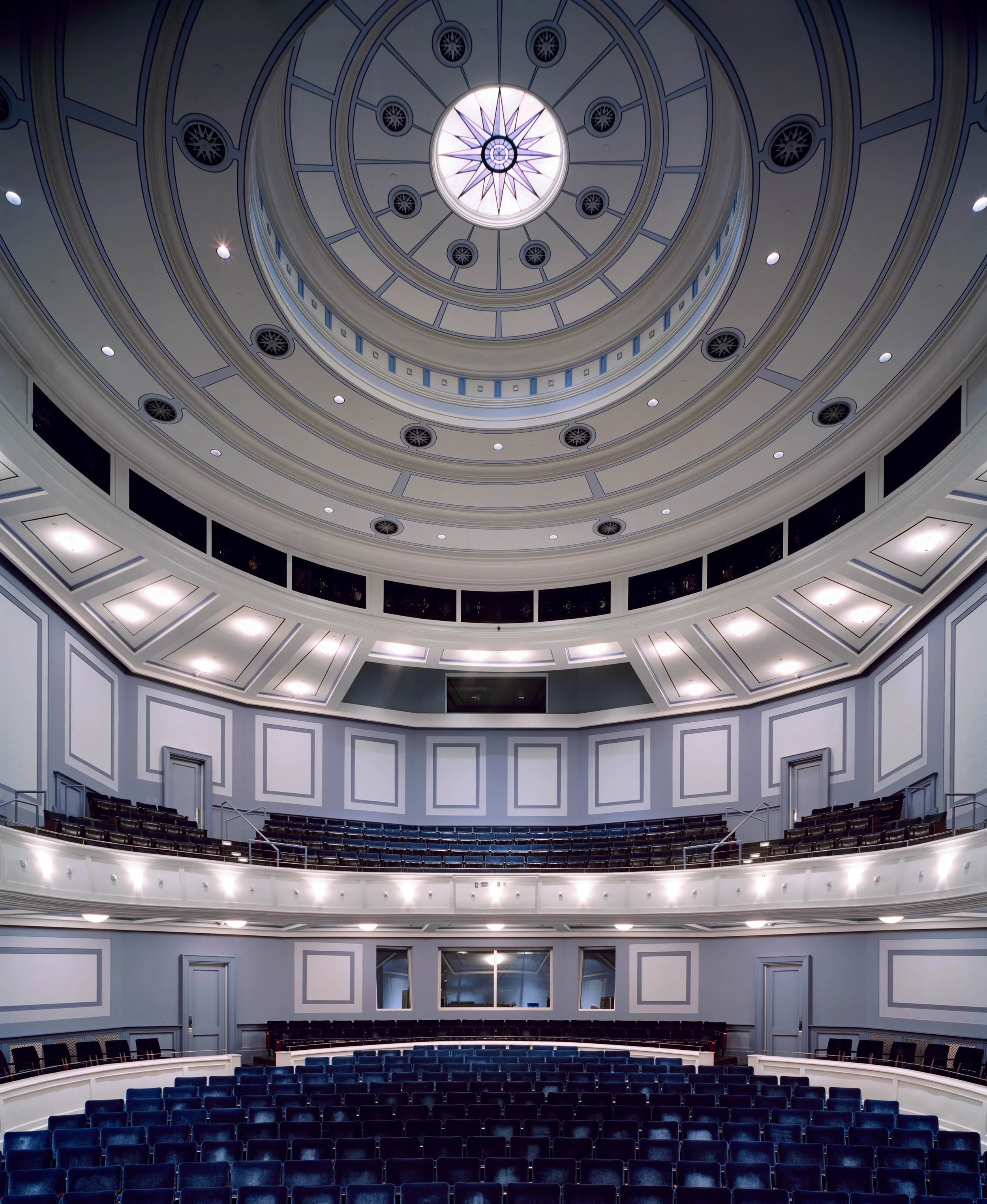 Interior view of a large, empty auditorium with rows of blue theater seats, multiple levels of balconies, and a domed ceiling with a decorative central light fixture.