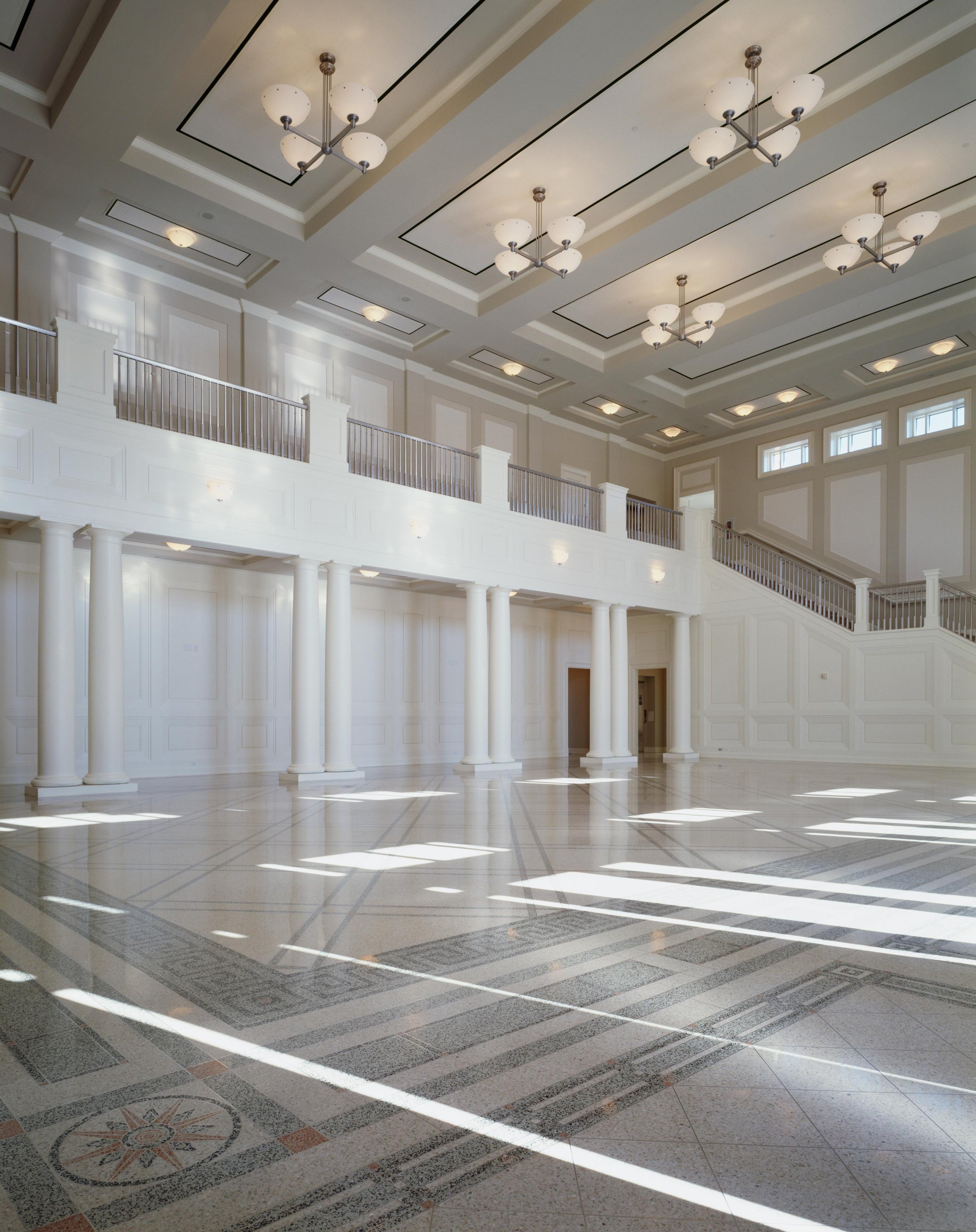 Large, bright lobby with white columns, ornate ceiling moldings, modern chandeliers, and sunlight streaming through windows creating striped shadows on the floor.