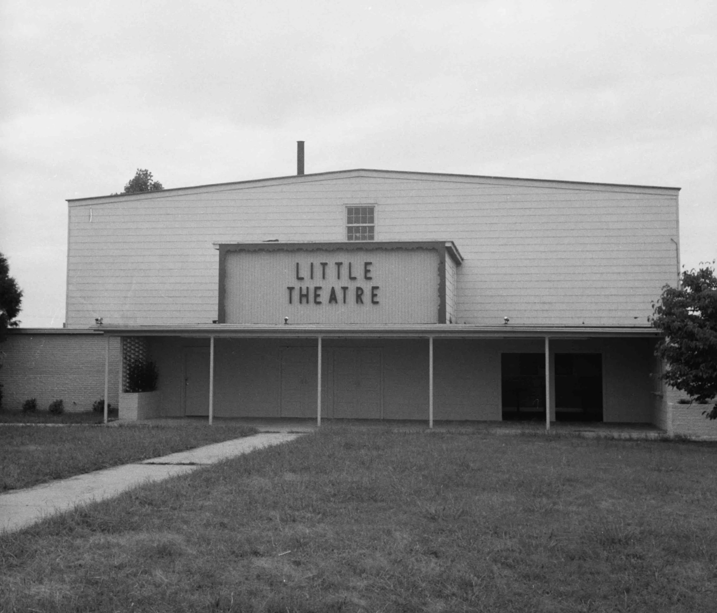Black and white photo of a building with a sign that reads "Little Theatre." The building has horizontal siding, a small window, and a porch area with support columns. Trees are visible in the background, and there is a sidewalk and well-maintained grass in the foreground.