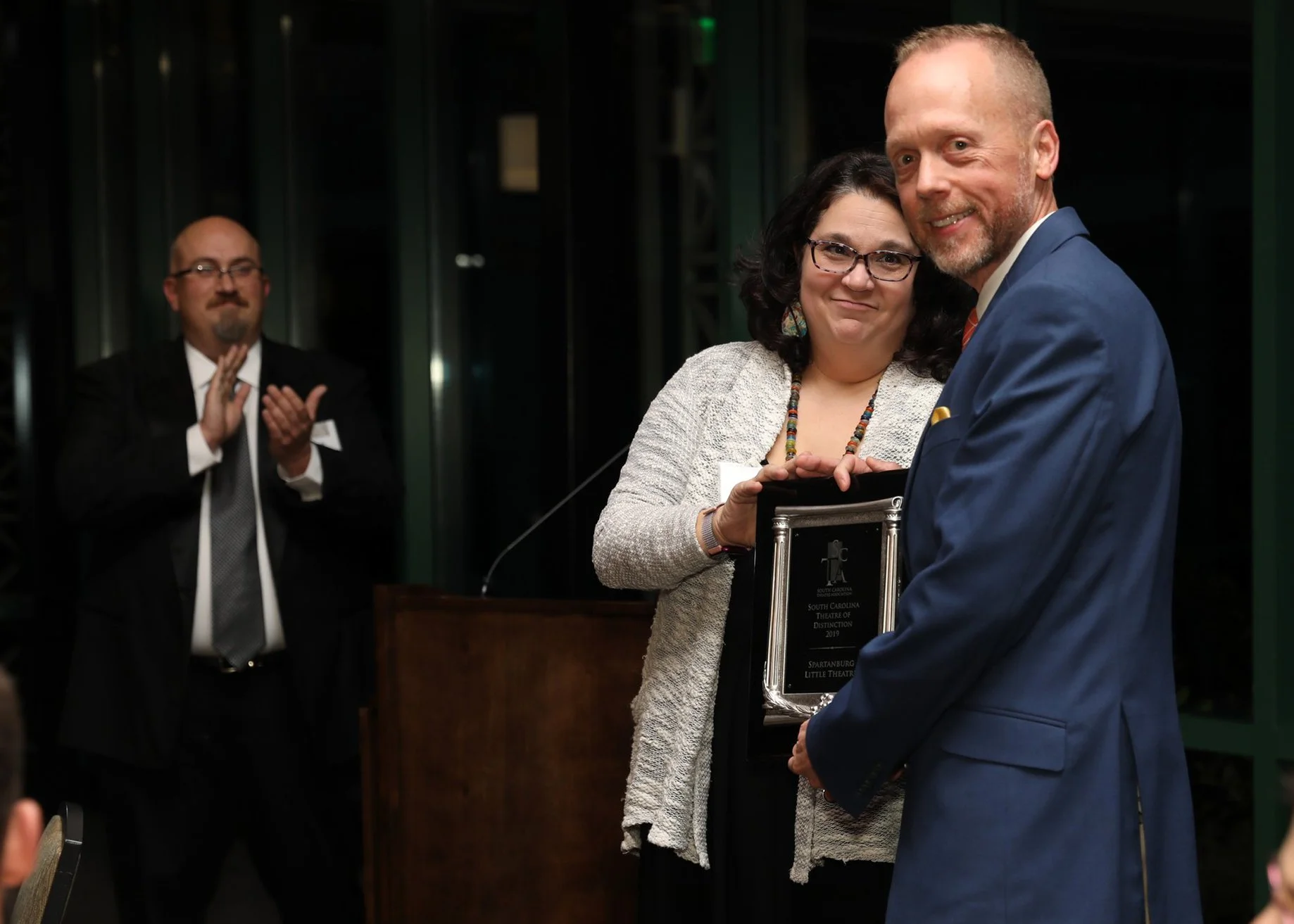 A man in a blue suit and a woman in glasses and a beige jacket are standing together at an award ceremony, holding an award plaque. Another man in the background is clapping, wearing a black suit and glasses.
