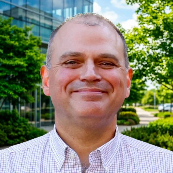 A smiling man with short hair in front of a building with glass windows, surrounded by green trees and shrubs.