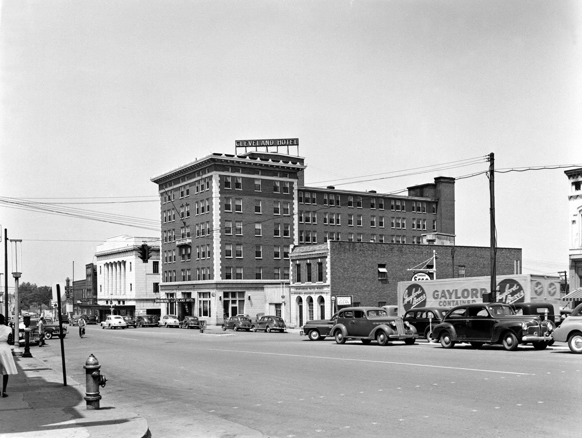 Black and white photo of a city street with vintage cars parked along the curb, a fire hydrant, pedestrians, and a large brick hotel building labeled 'Cleveland Hotel' in the background.