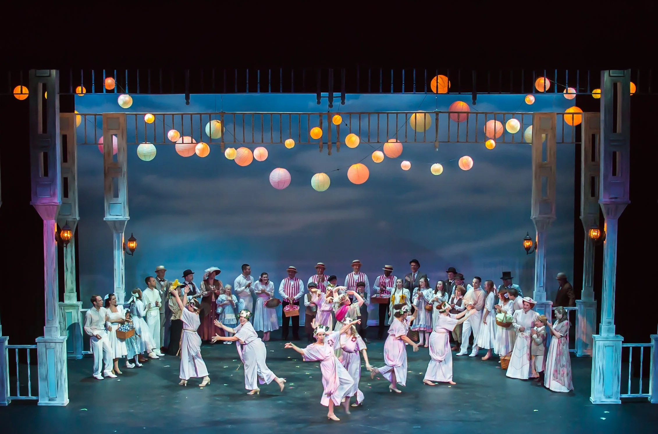 On stage, a large group of performers dressed in historical costumes, with some dancing and others standing in a line, under a decorated sky with hanging paper lanterns in various colors.