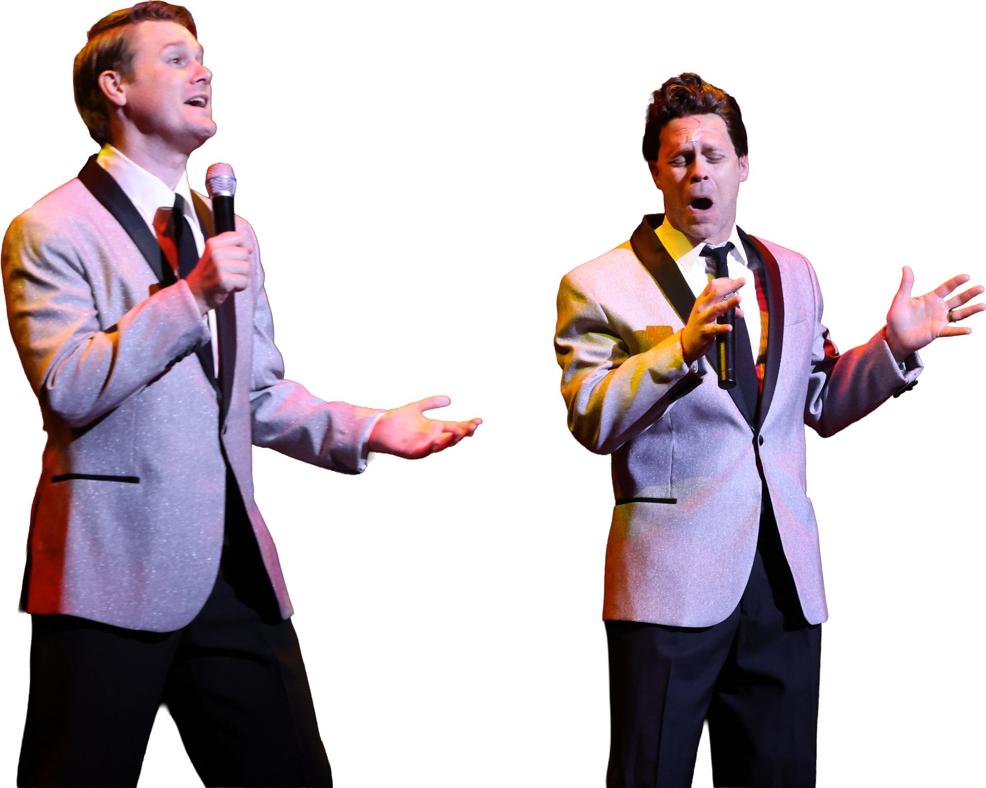 Two men on stage, both wearing silver jackets with black lapels and ties, holding microphones, engaged in a singing performance with expressive gestures and facial expressions.