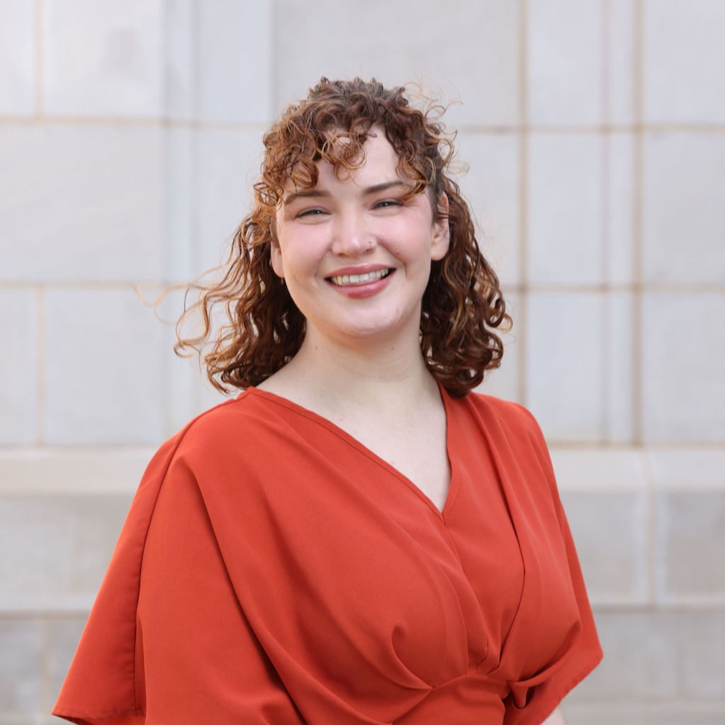 A woman with curly red hair and a nose piercing, wearing an orange top, smiling outdoors in front of a beige tiled wall.