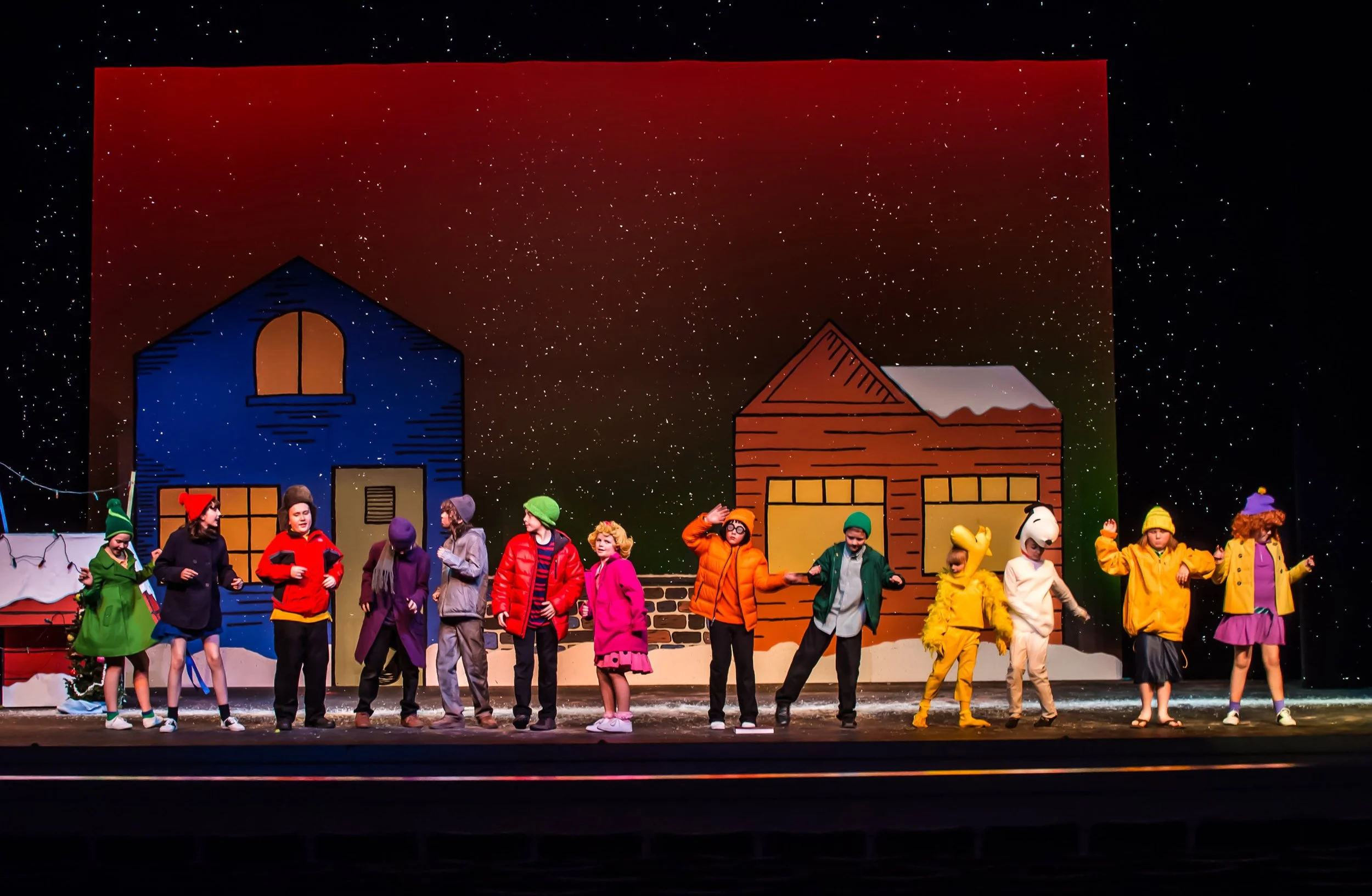 Children performing a play on stage with winter holiday decorations, painted houses, and a snowy backdrop.