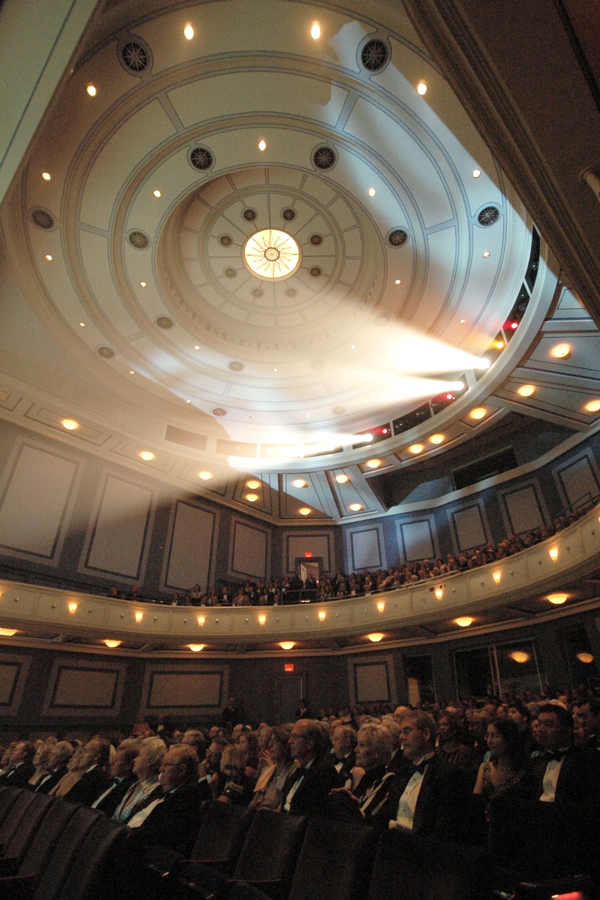 Interior of a large auditorium filled with seated audience members in formal attire, with multiple balconies and a ornate ceiling with circular light fixtures.