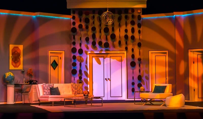 A stage set resembling a mid-century modern living room with colorful decor and dramatic lighting, including a white sofa, a yellow chair, a coffee table, patterned pillows, and wall art, with a door and a hanging chandelier.