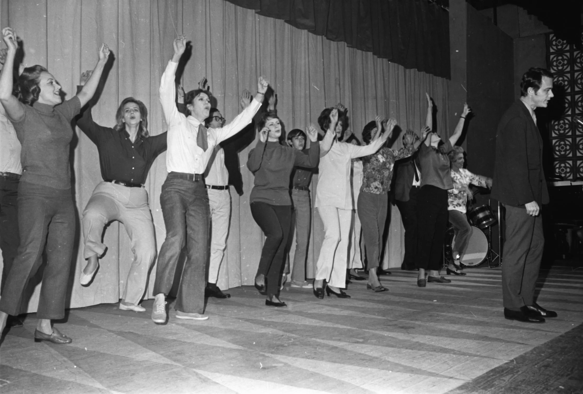 A black and white photo of a group of people, possibly performers, holding hands and raising their arms on stage with a curtain backdrop. A man at the front is walking away from the group.