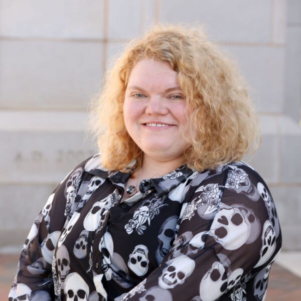 A woman with curly blonde hair smiling outdoors, wearing a black shirt with white skull patterns.